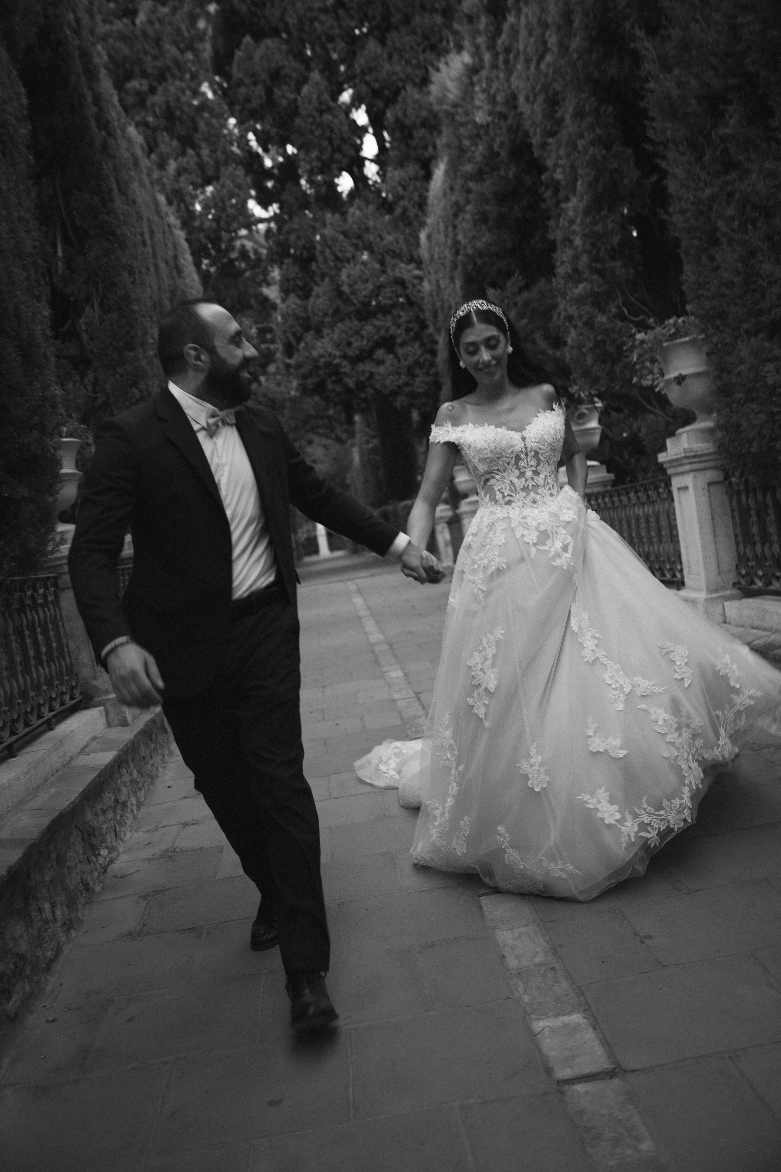 Pareja de novios bailando en una calle entre árboles, ella lleva vestido de novia con detalles de encaje y él lleva traje oscuro con pajarita.