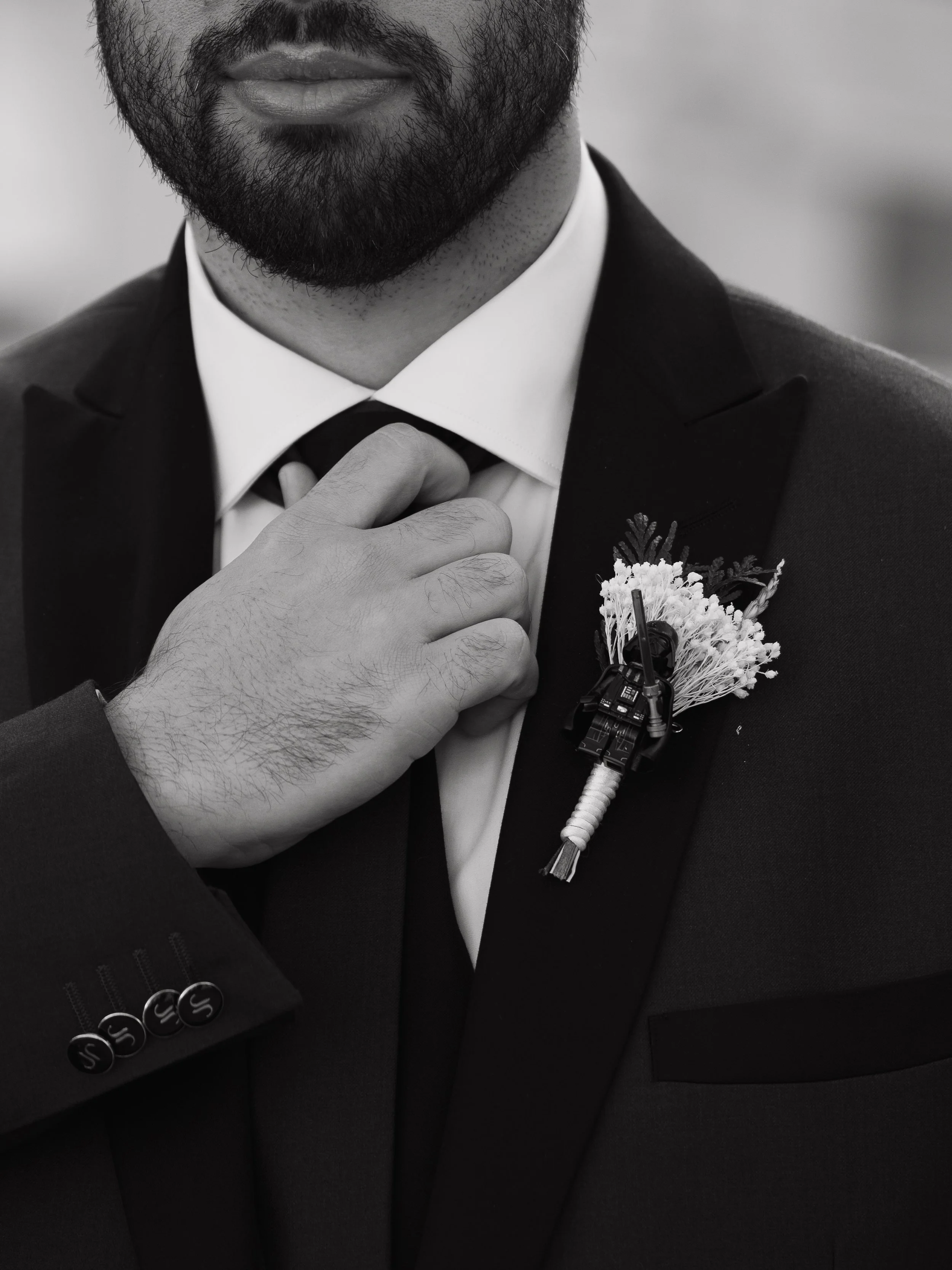 Hombre con barba ajustando su corbata, usando traje formal, con una flor y un micrófono en el saco. La imagen es en blanco y negro.