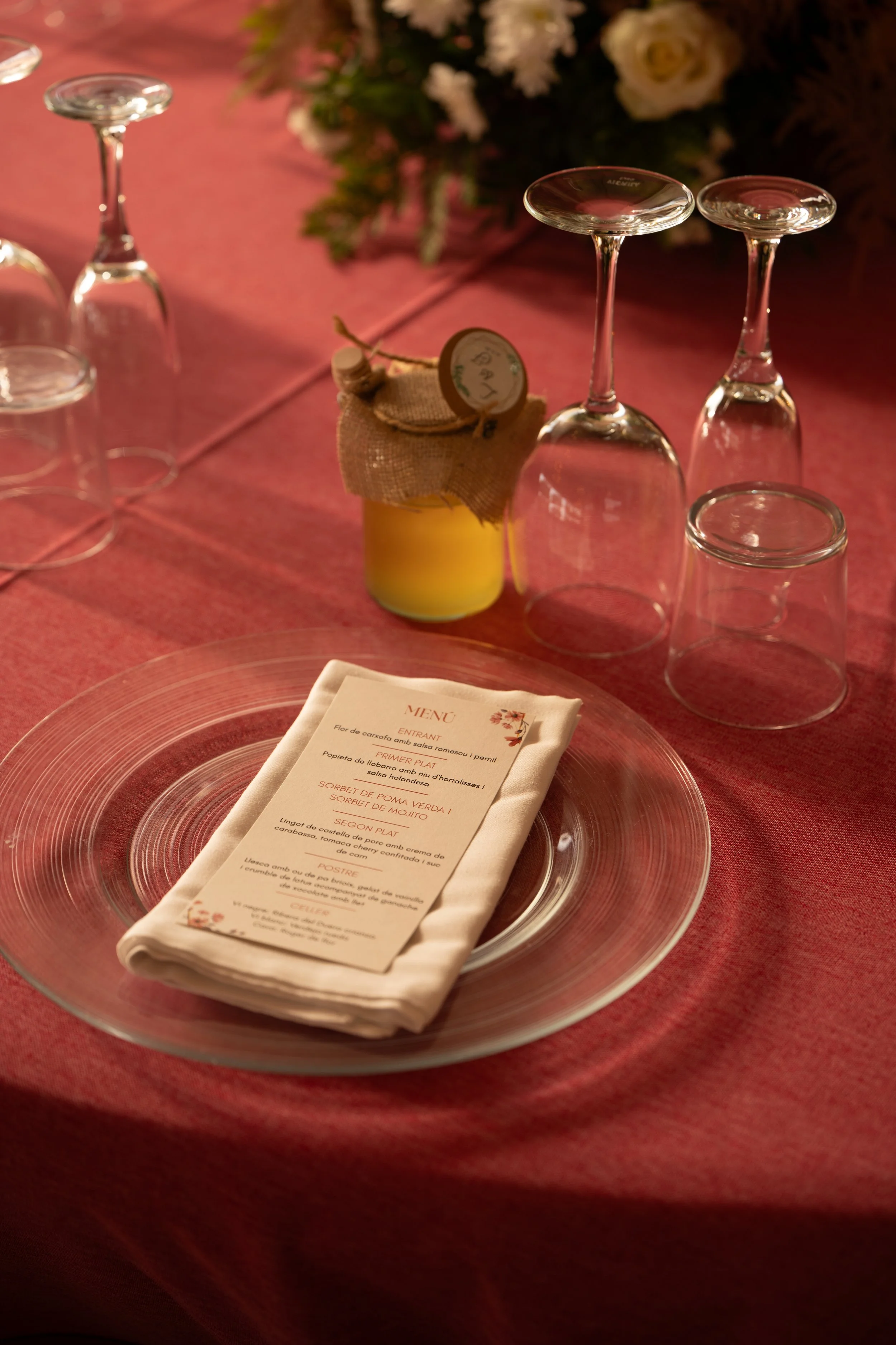 Vasos de vino y un florero con flores en una mesa con manteles rosas, además de un plato con un menú y un cubierto, en un ambiente elegante.