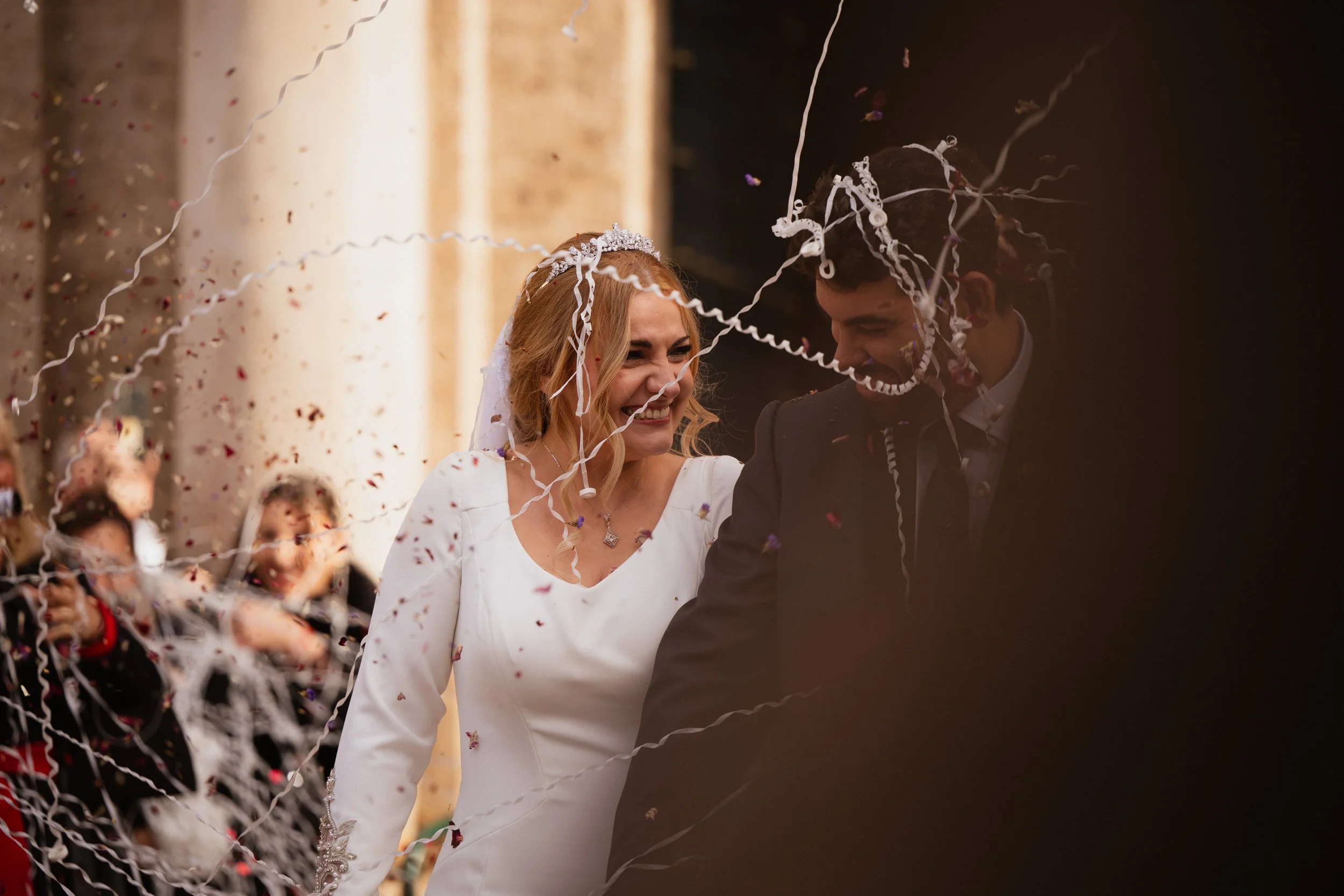 Pareja casándose, mujer con vestido blanco y tiara, hombre con traje, rodeados de confeti y serpentinas, celebrando en ceremonia de boda.