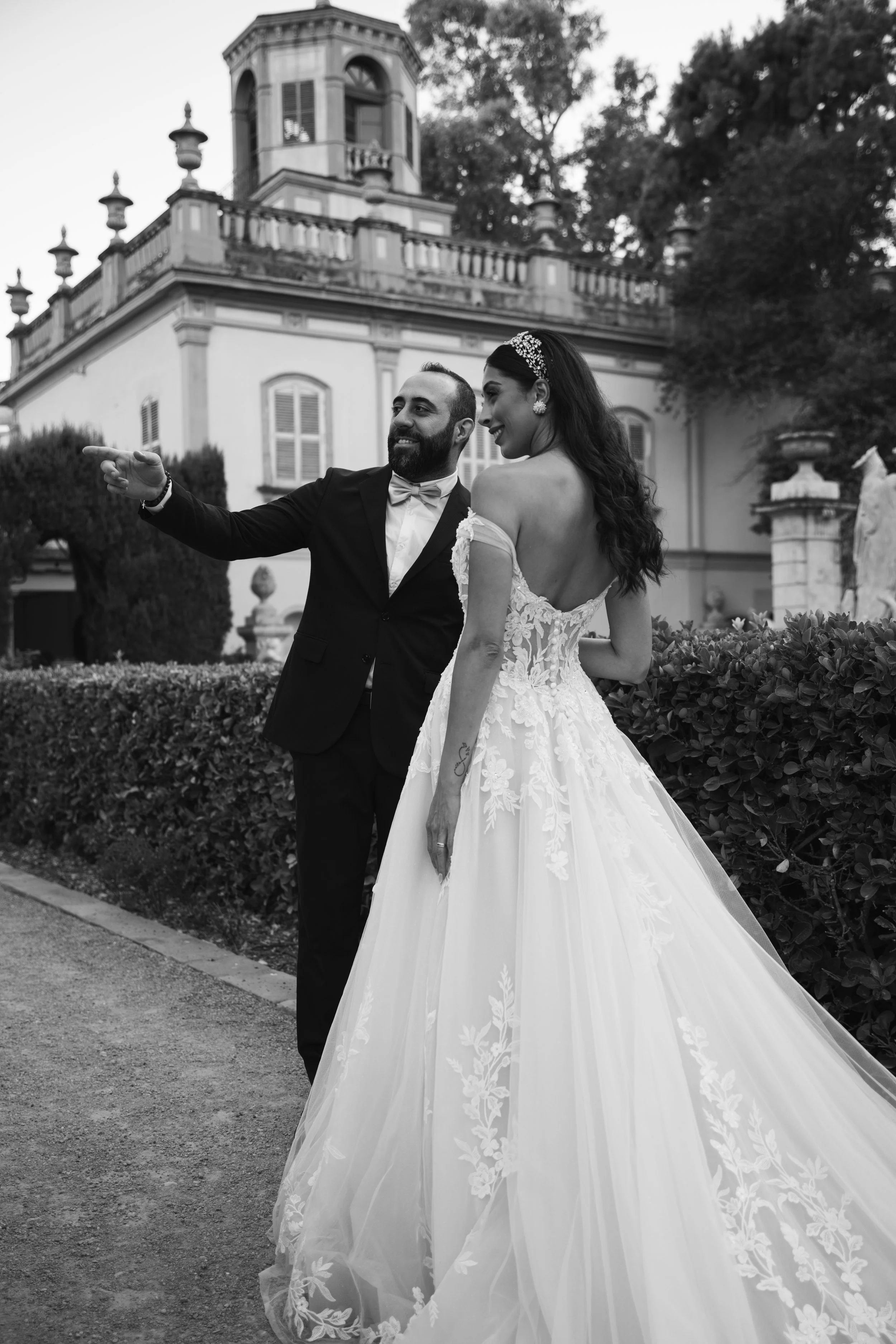Un hombre y una mujer en vestido de boda sonriendo juntos, con un edificio antiguo y árboles de fondo, en una ceremonia nupcial al aire libre en color sepia.