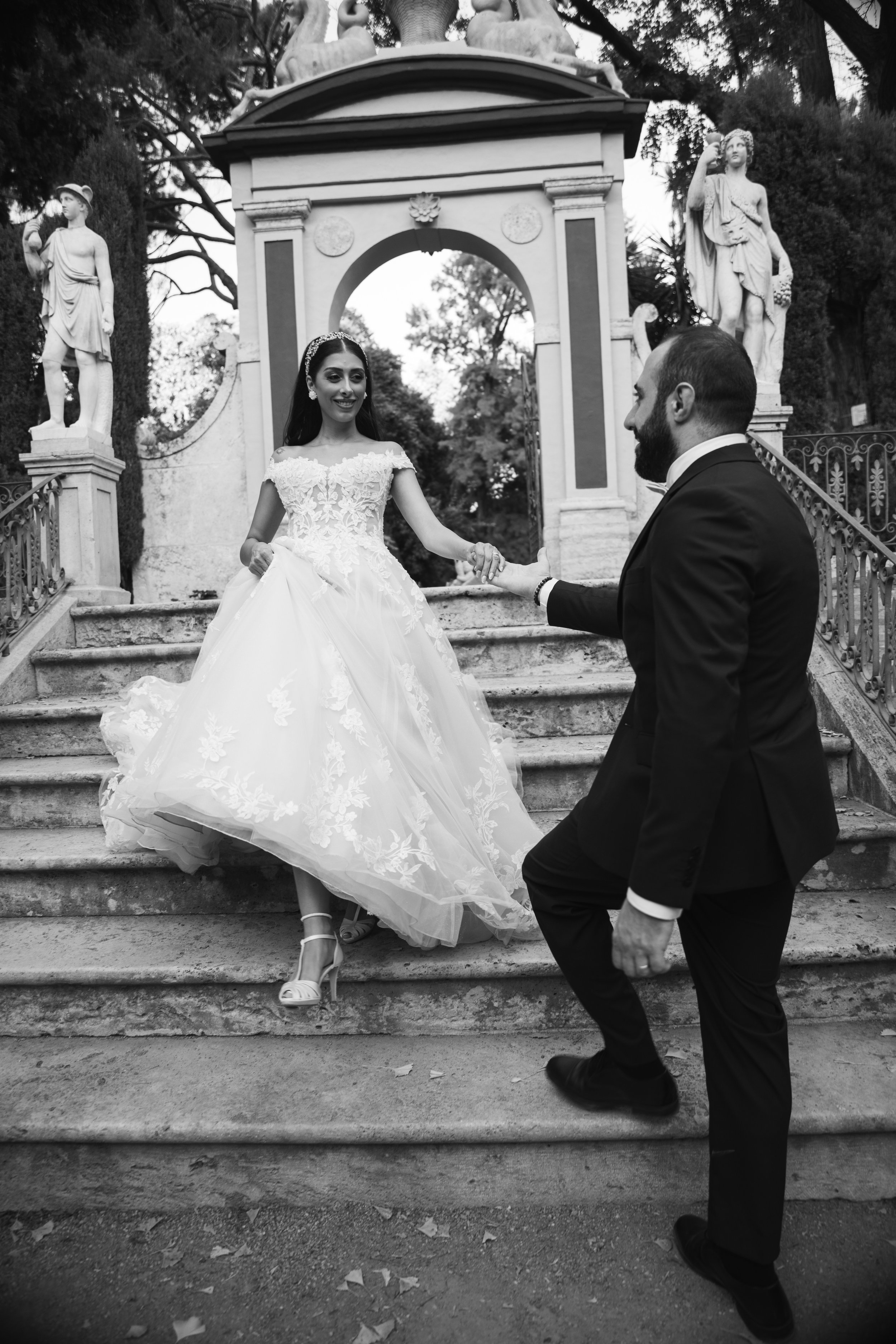 Una pareja de novios bailando en un escenario de piedra, la novia lleva un vestido de novia largo y el novio lleva un traje negro. Están rodeados de esculturas clásicas y un arco en un entorno externo con árboles en el fondo.