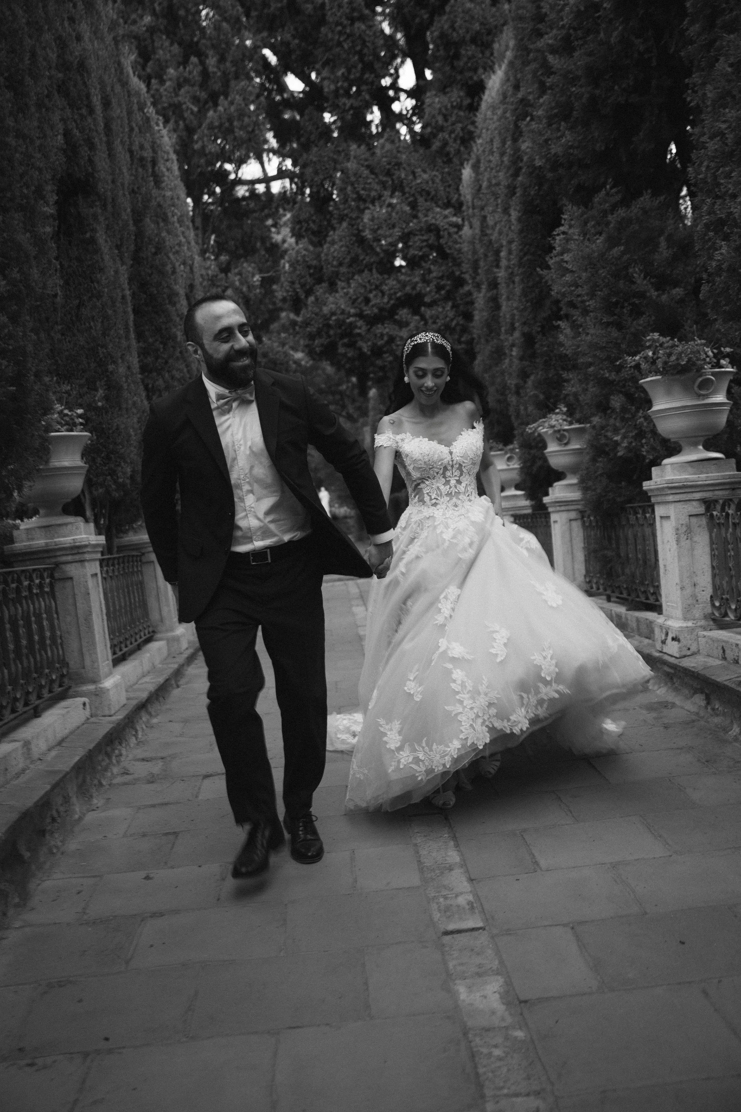 Pareja de novios bailando en una calle, ella vestida con un vestido de novia y él con esmoquin, rodeados de árboles y una barandilla de piedra.