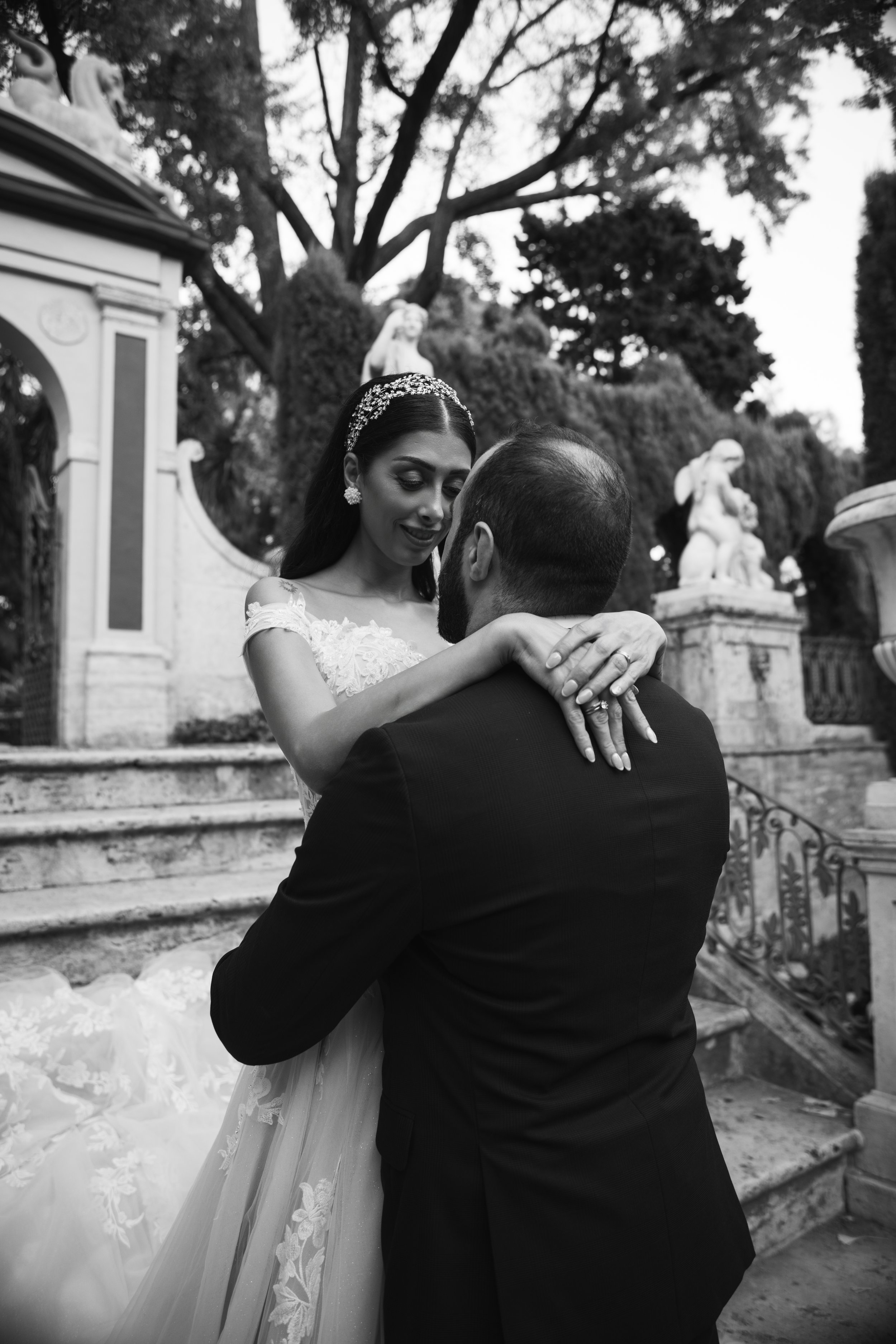 Una pareja en un abrazo, con la mujer vestida de novia y el hombre de traje, en un entorno exterior con escaleras y estatuas clásicas, en un día soleado.