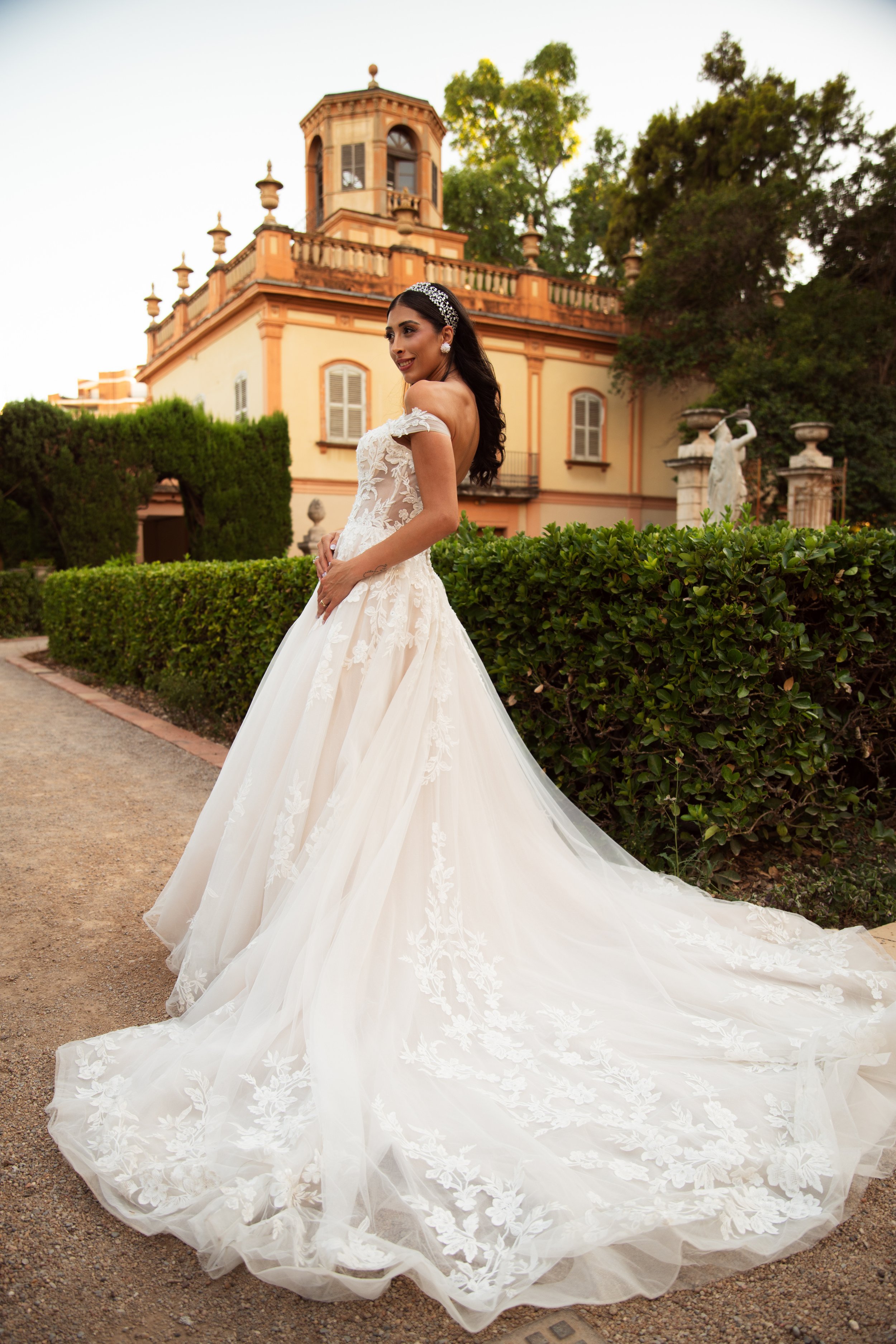 Mujer en vestido de novia con velo y accesorios, posando en jardín frente a edificio antiguo con estatuas y árboles verdes.