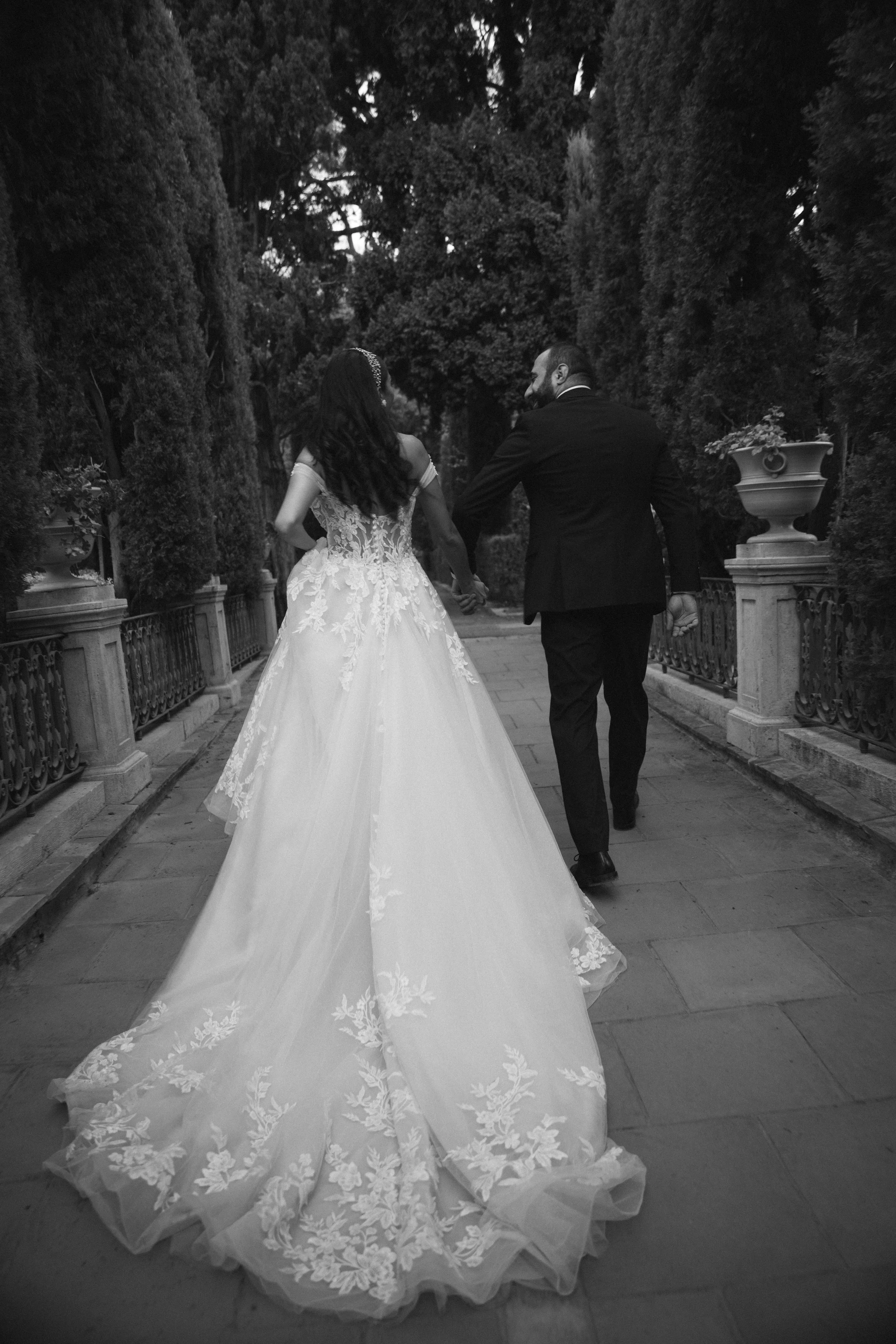 Pareja de novios caminando de la mano en un camino flanqueado por arbustos y macetas, vista desde atrás. Ella lleva vestido de novia con cola larga de encaje floral, él lleva saco negro y pantalón oscuro.