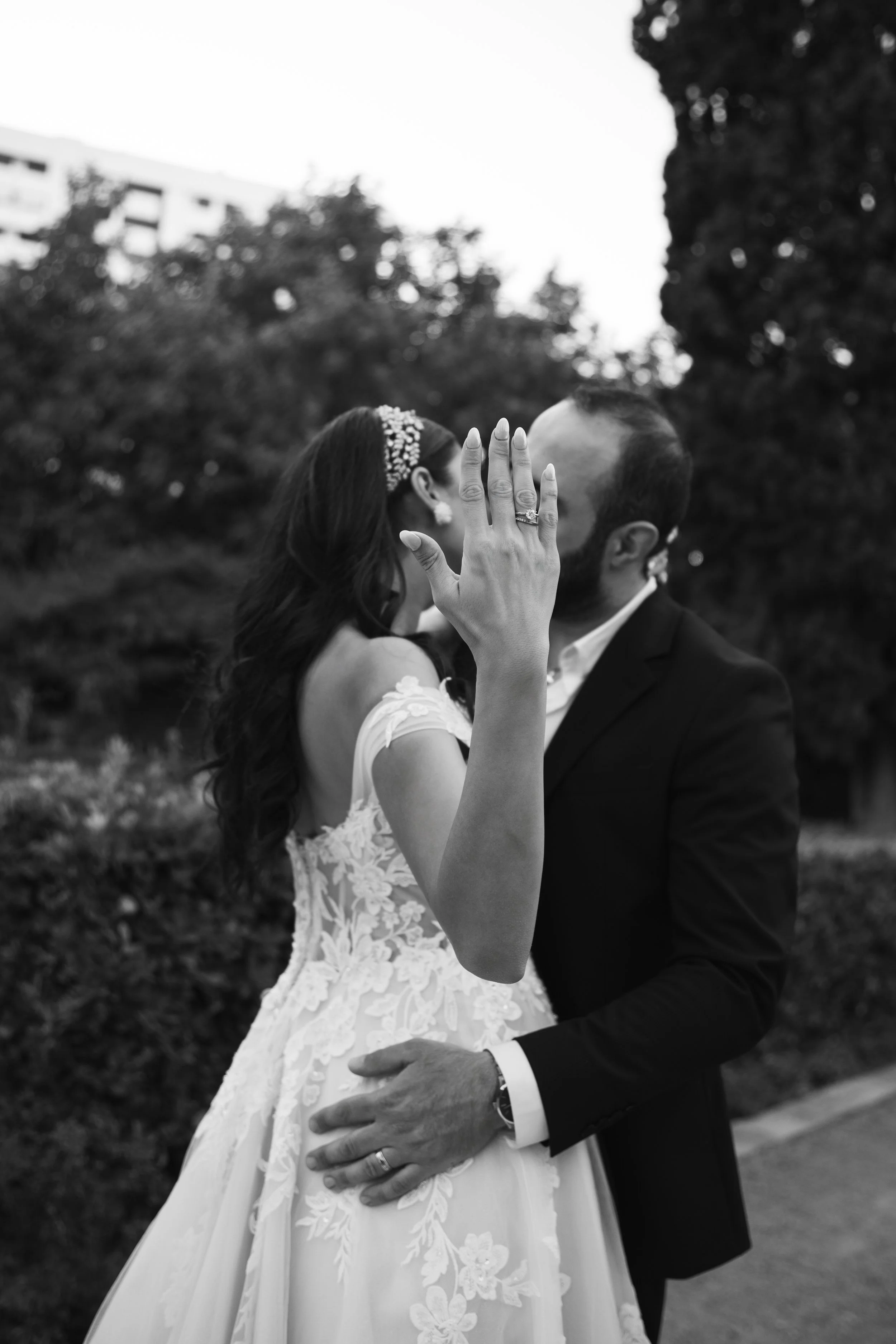 Pareja de novios en boda, la novia usa vestido de encaje y diadema, el novio en traje oscuro, en fondo natural con árboles.