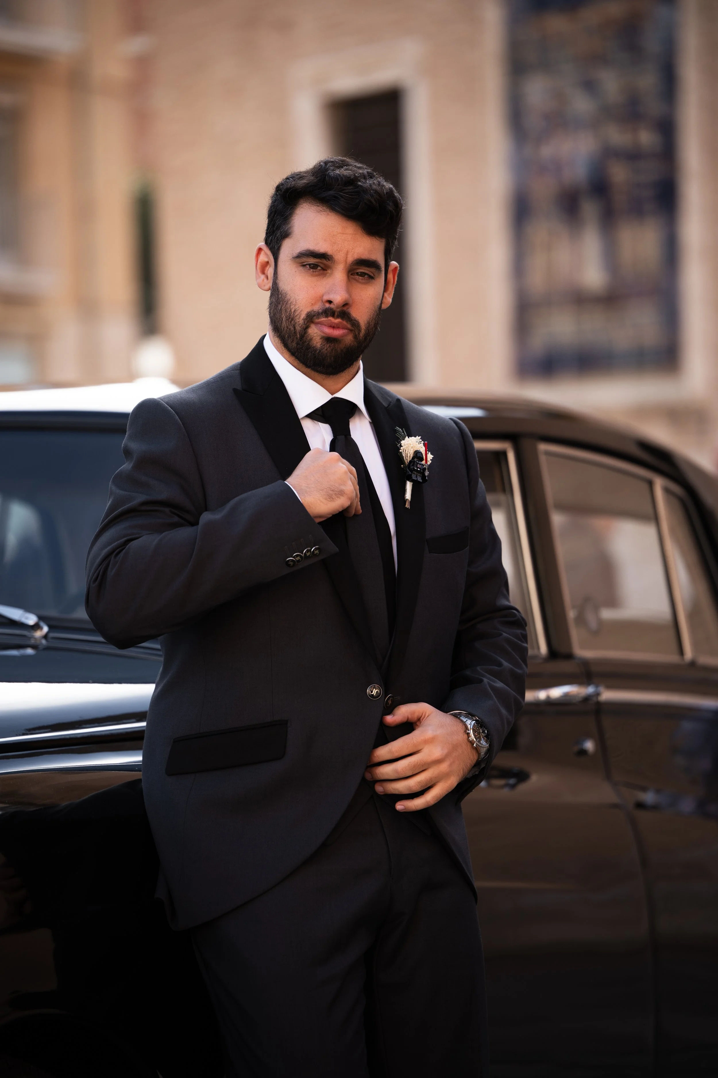 Hombre con barba y cabello oscuro usando traje negro, corbata y reloj, posando junto a un automóvil negro en exteriores urbanos.