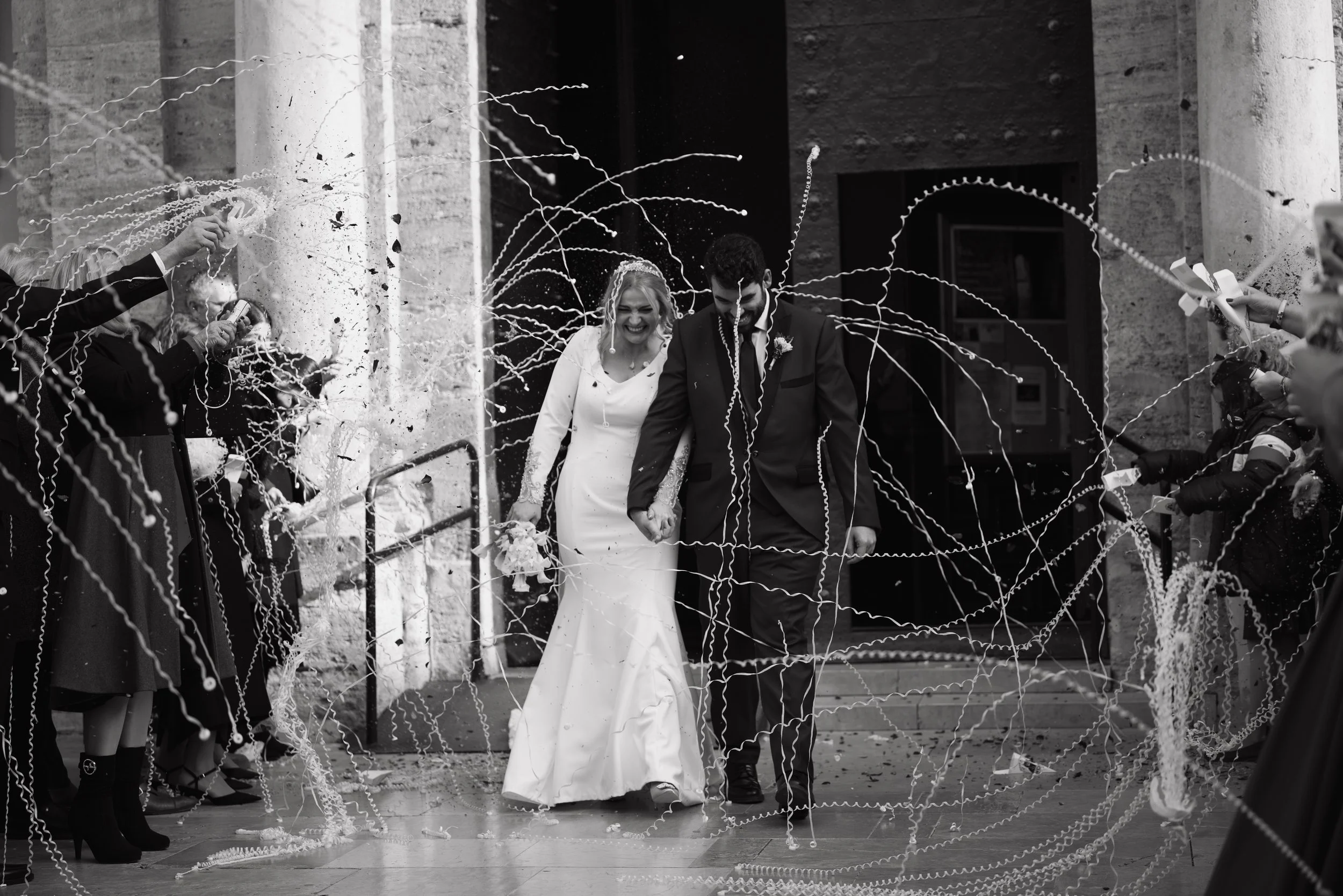 Una pareja de novia y novio caminando de la mano en una boda, rodeados de invitados lanzando serpentinas en un entorno exterior con columnas de piedra.