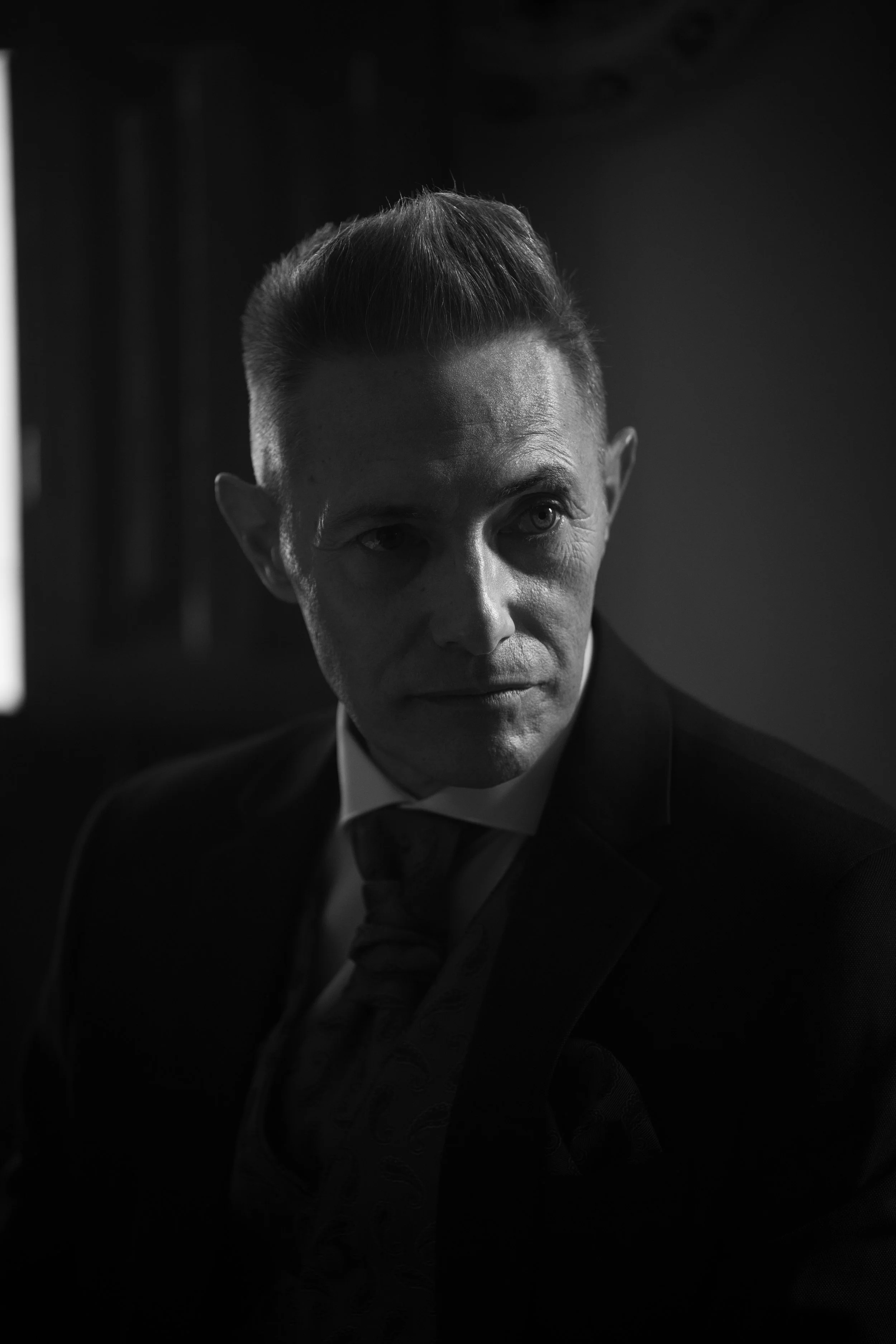 Hombre con cabello peinado hacia atrás, usando traje y corbata, en un entorno oscuro.