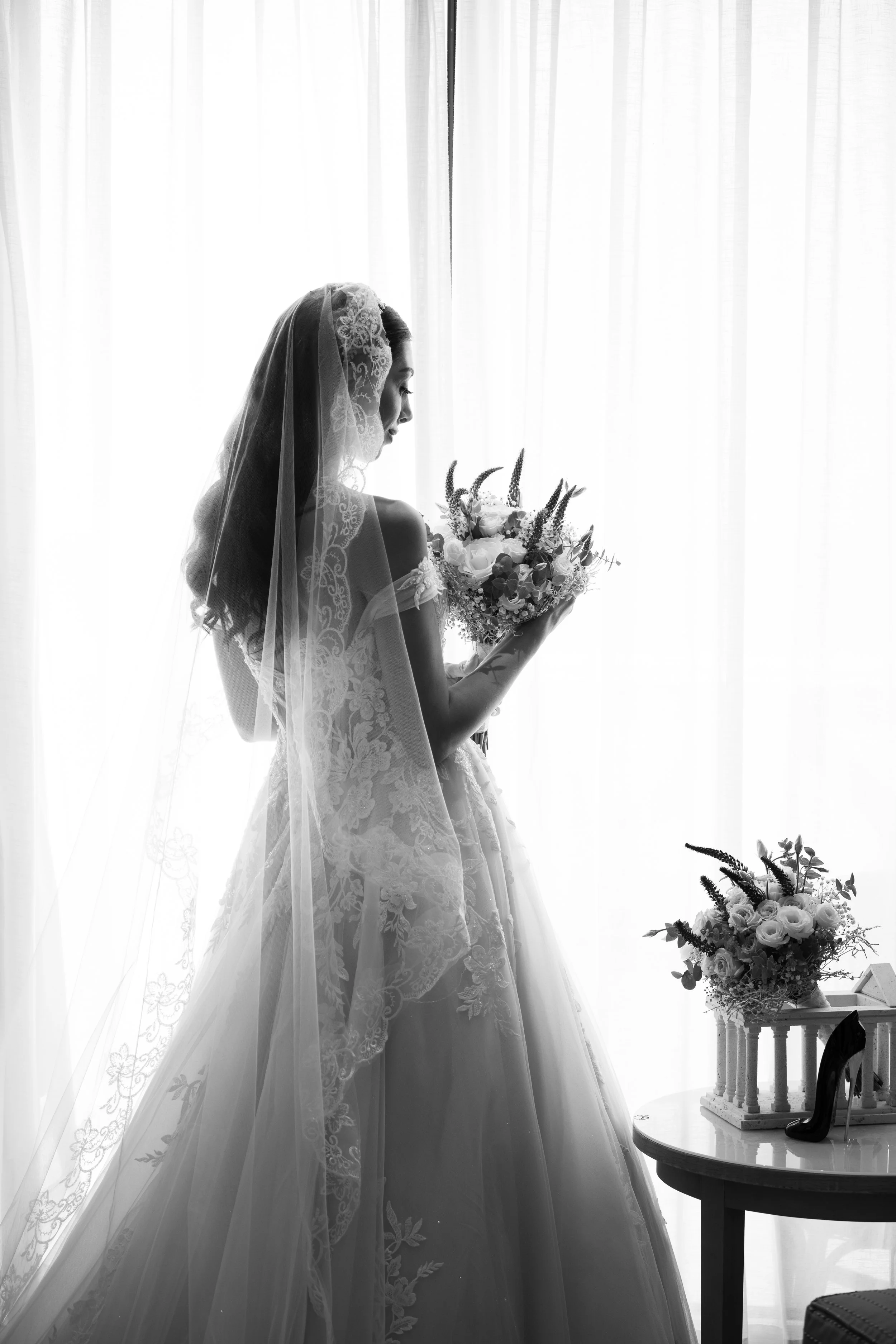 Mujer con vestido de novia y velo, sosteniendo un ramo de flores, de espaldas, en un ambiente con cortinas de fondo, en blanco y negro.