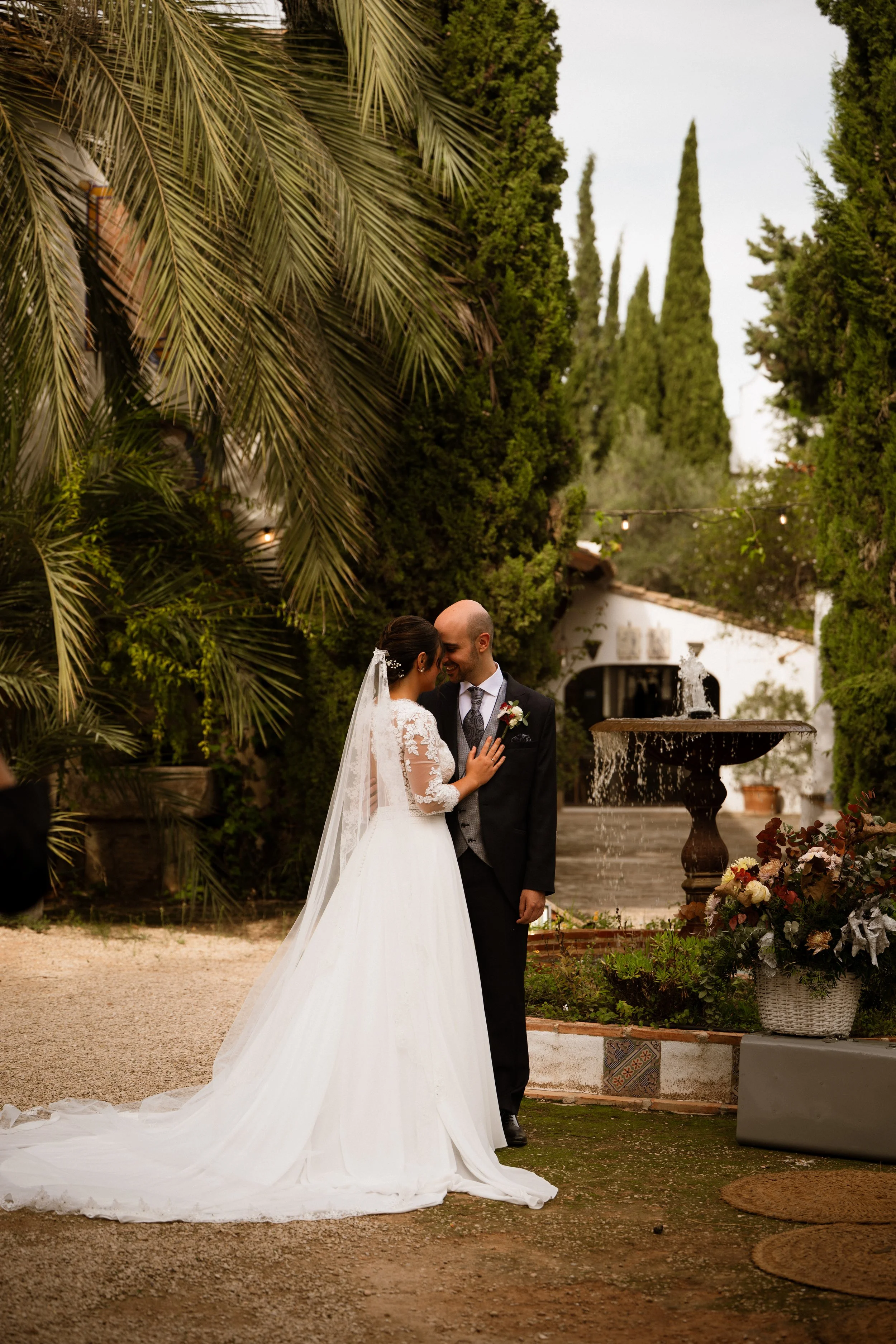 Pareja de recién casados en un jardín, la novia viste un vestido largo blanco con velo y el novio lleva traje negro, rodeados de vegetación y decoraciones florales.
