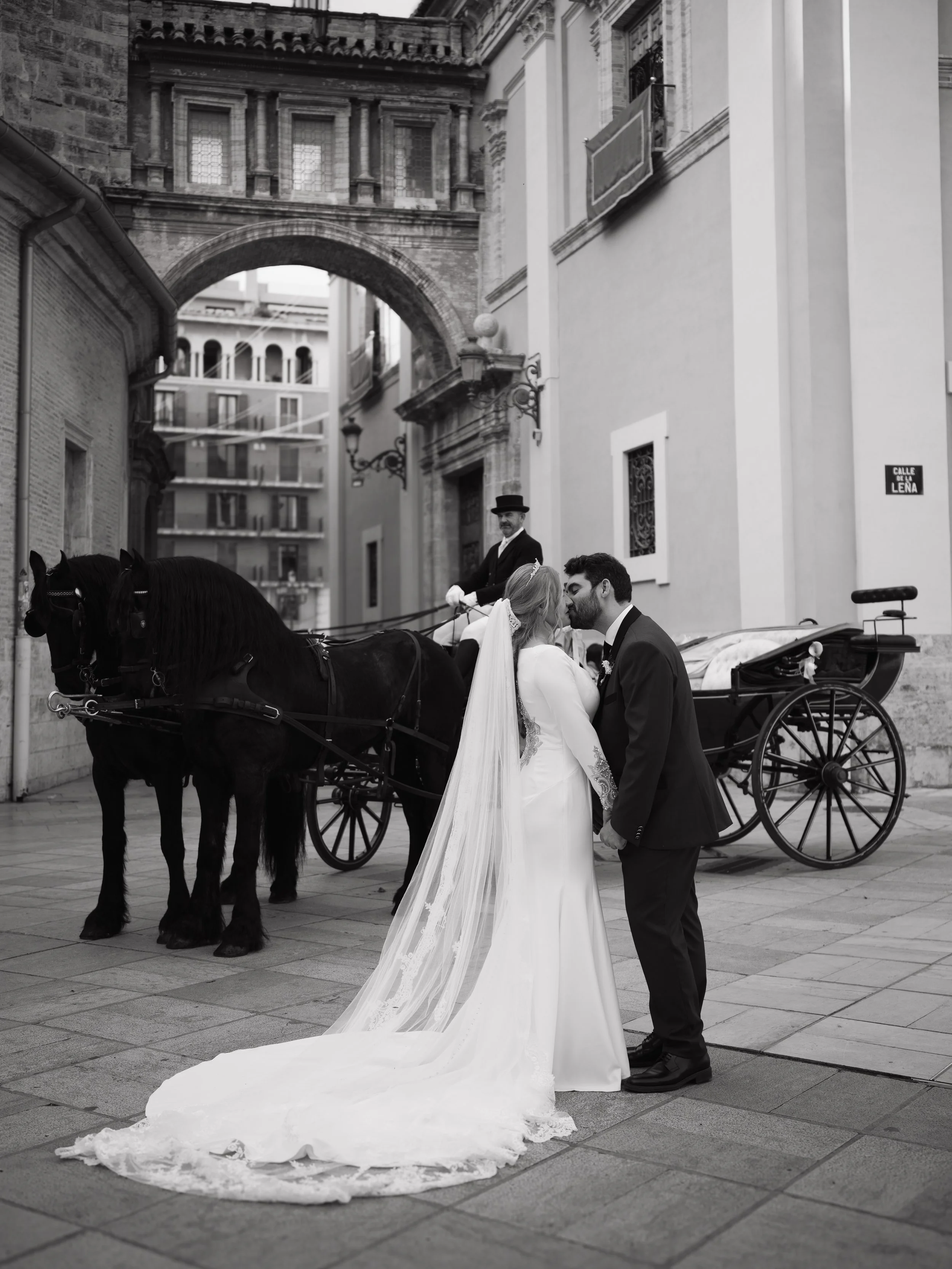 Boda en la calle con un coche de caballos, una pareja de novios que se besan, una persona en el carruaje y edificios históricos de fondo.