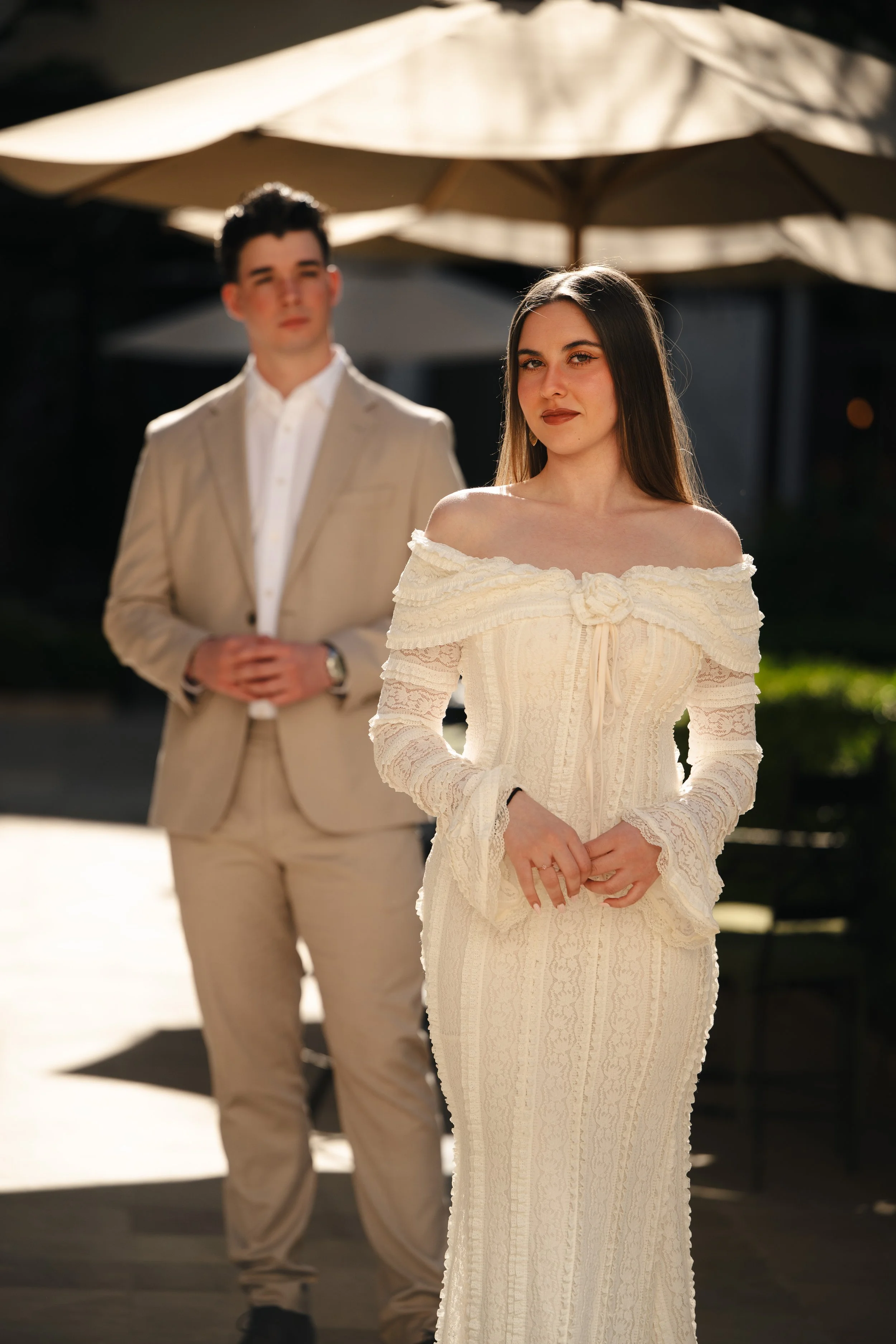 Una pareja con ropa formal en un entorno exterior, la mujer en primer plano con un vestido largo y la máscara de la mujer, el hombre en el fondo con traje claro y camisa blanca, debajo de una sombrilla beige en un día soleado.