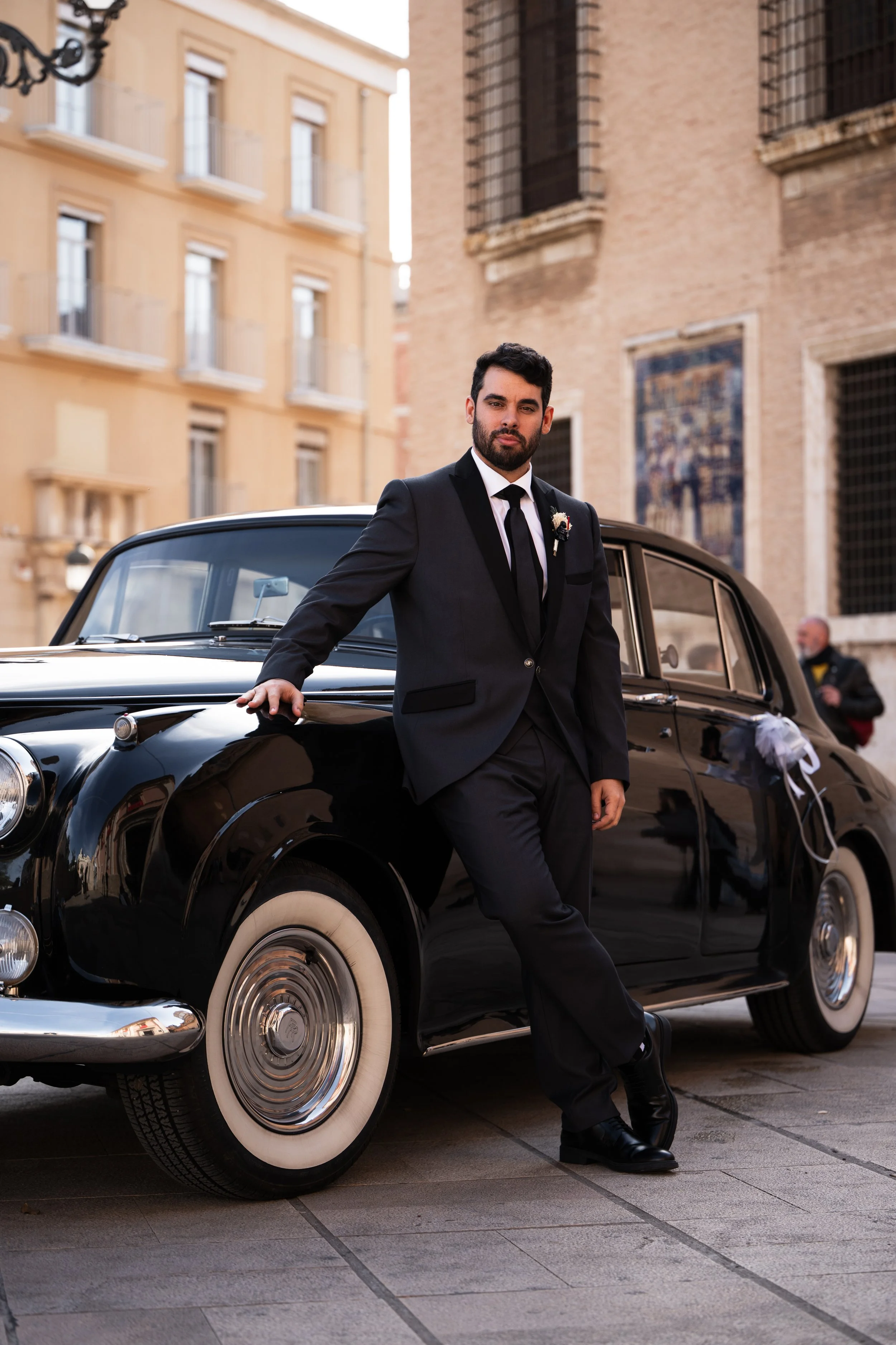 Hombre vestido con traje negro y corbata, apoyado en un coche clásico negro con ruedas blancas, en un entorno urbano con edificios antiguos y murales en el fondo.