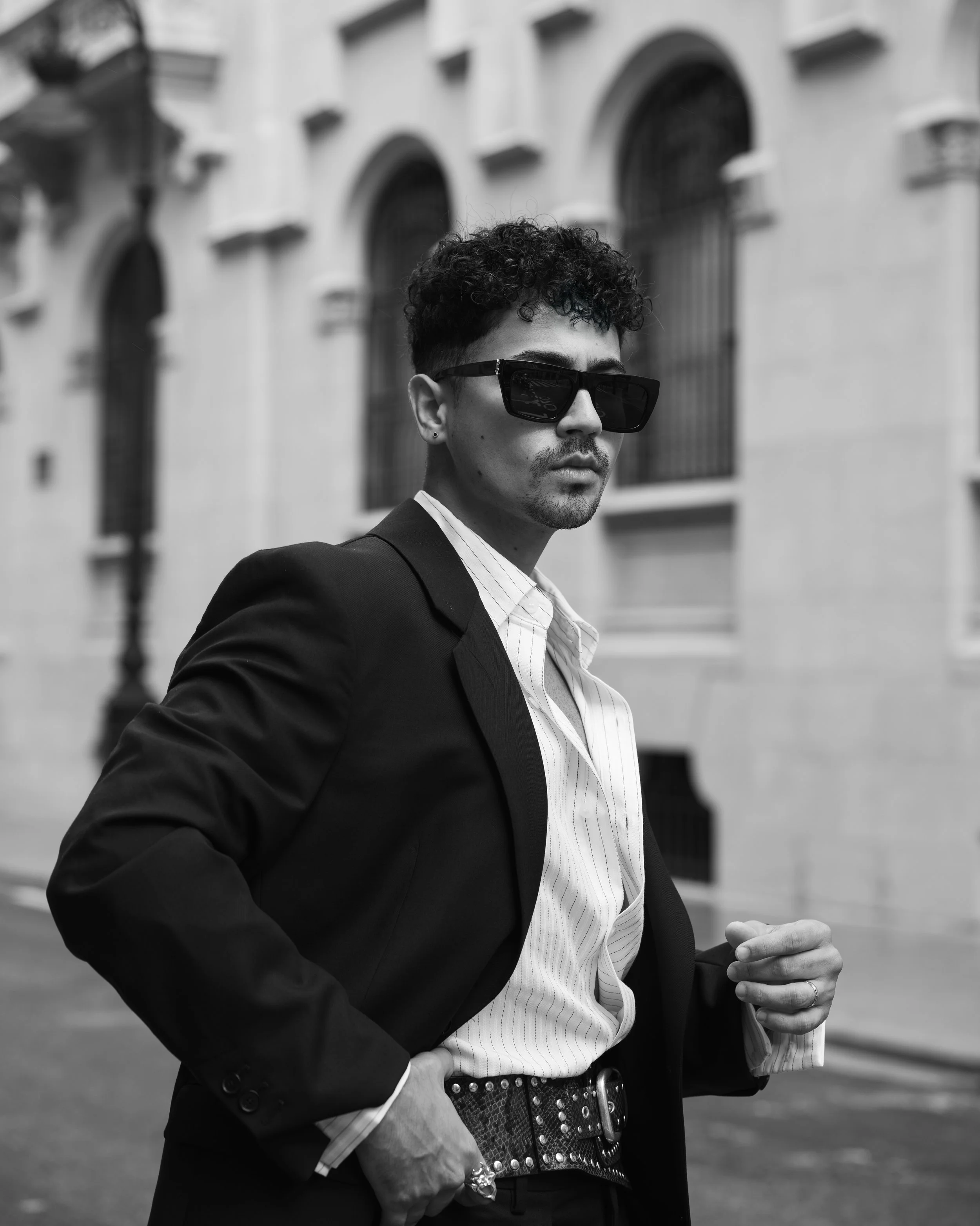 Hombre con cabello rizado y gafas oscuras viste traje negro y camisa blanca, en una calle con edificios de fondo y en blanco y negro.