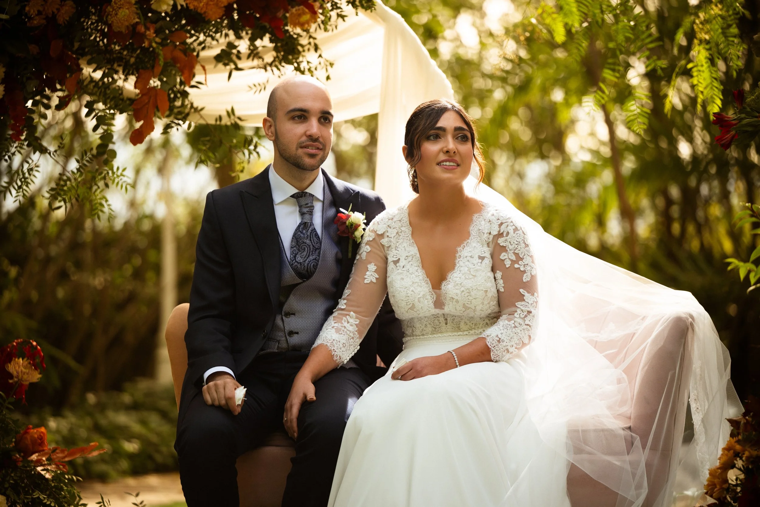 Una pareja de novios en una boda al aire libre, sentados juntos en un entorno con vegetación y flores, con la novia vestida con un vestido de encaje y el novio con traje formal.