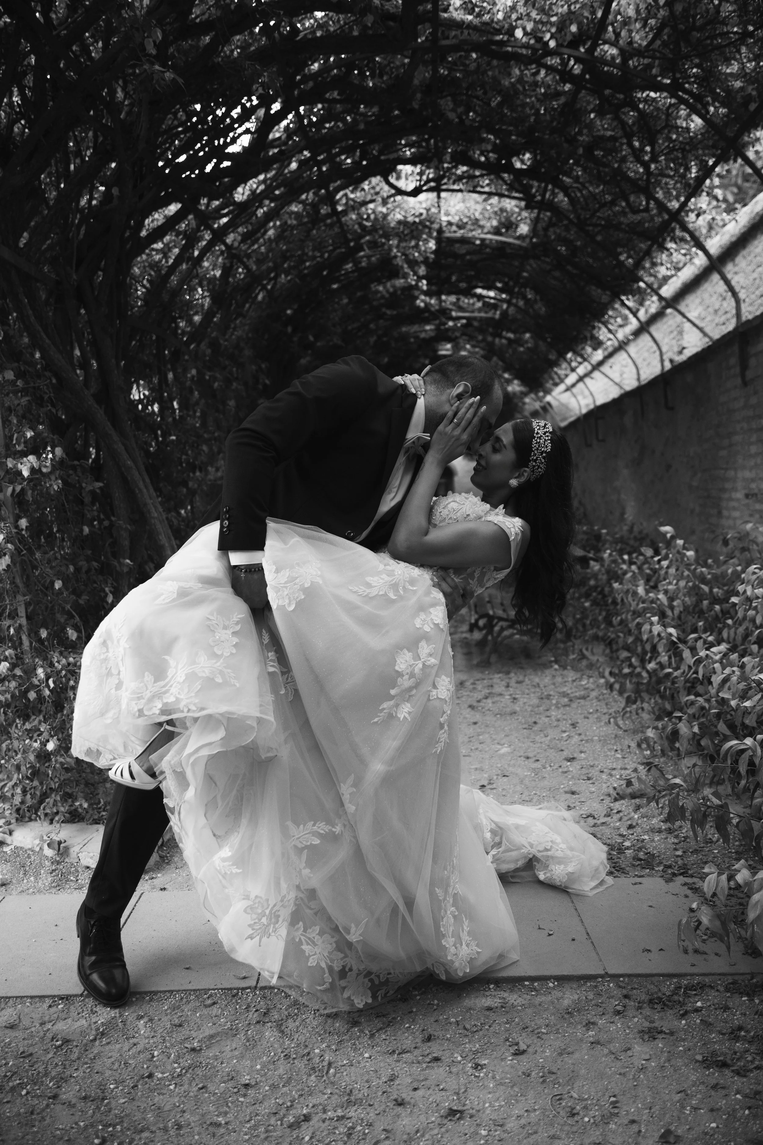 Una pareja de novios en una boda, el hombre vestido con traje negro y la mujer con un vestido de novia con encaje y diadema, en un pasaje arbolado