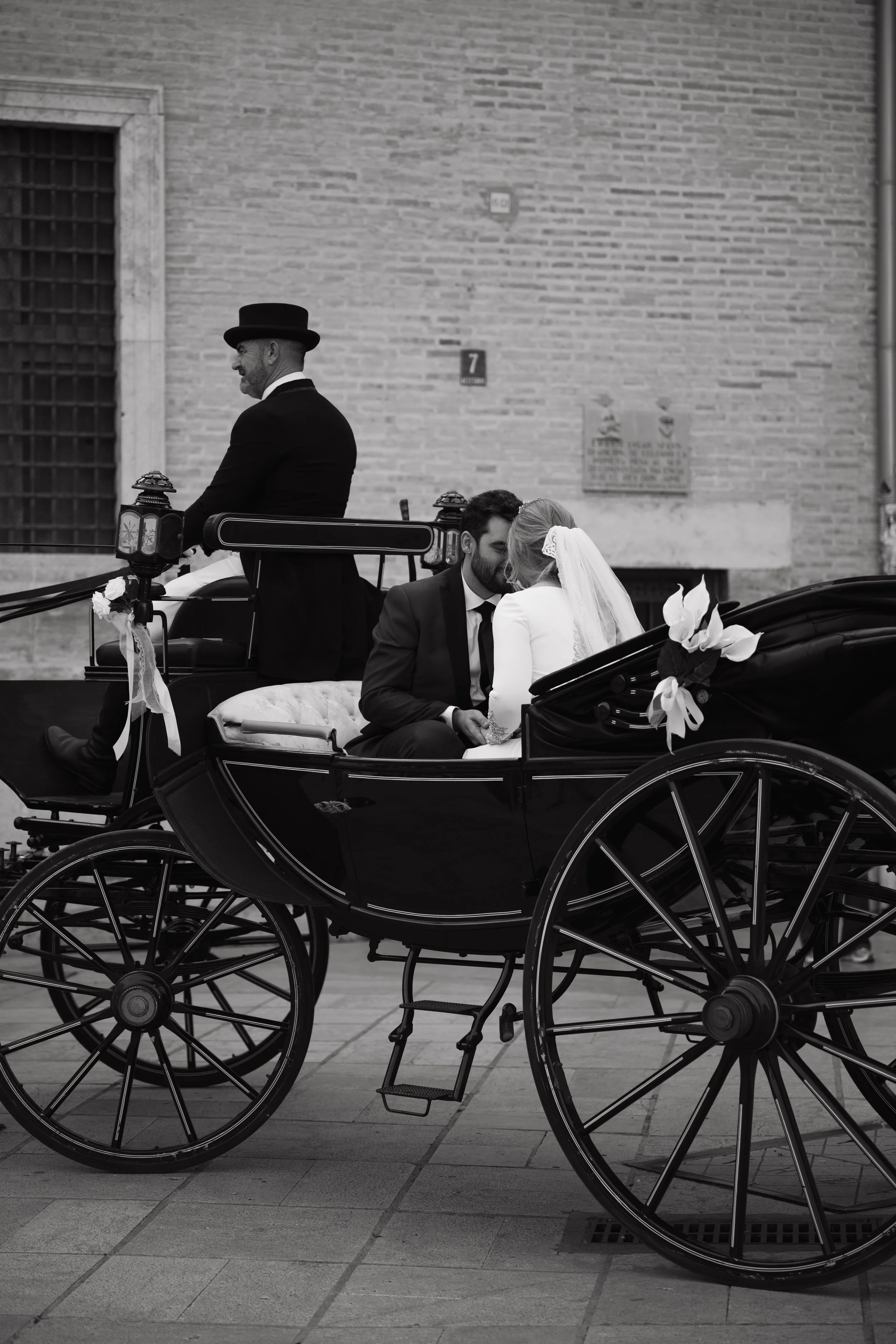 Dos personas en un carruaje de caballos, un hombre y una mujer en la parte trasera, la mujer usa un velo y el hombre usa un traje formal, con una pared de ladrillos en el fondo.
