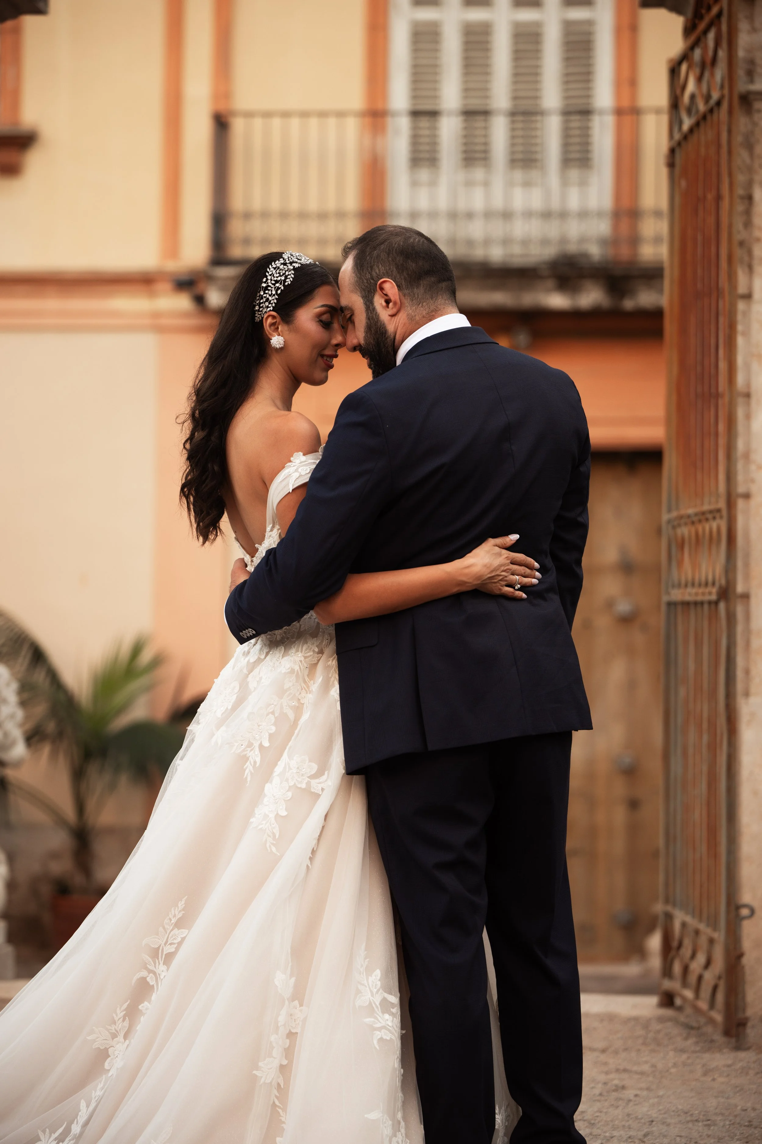 Pareja de novios en un abrazo, la mujer con vestido de boda y accesorios brillantes, el hombre con traje oscuro, en un entorno exterior con paredes de colores cálidos y una puerta de madera.
