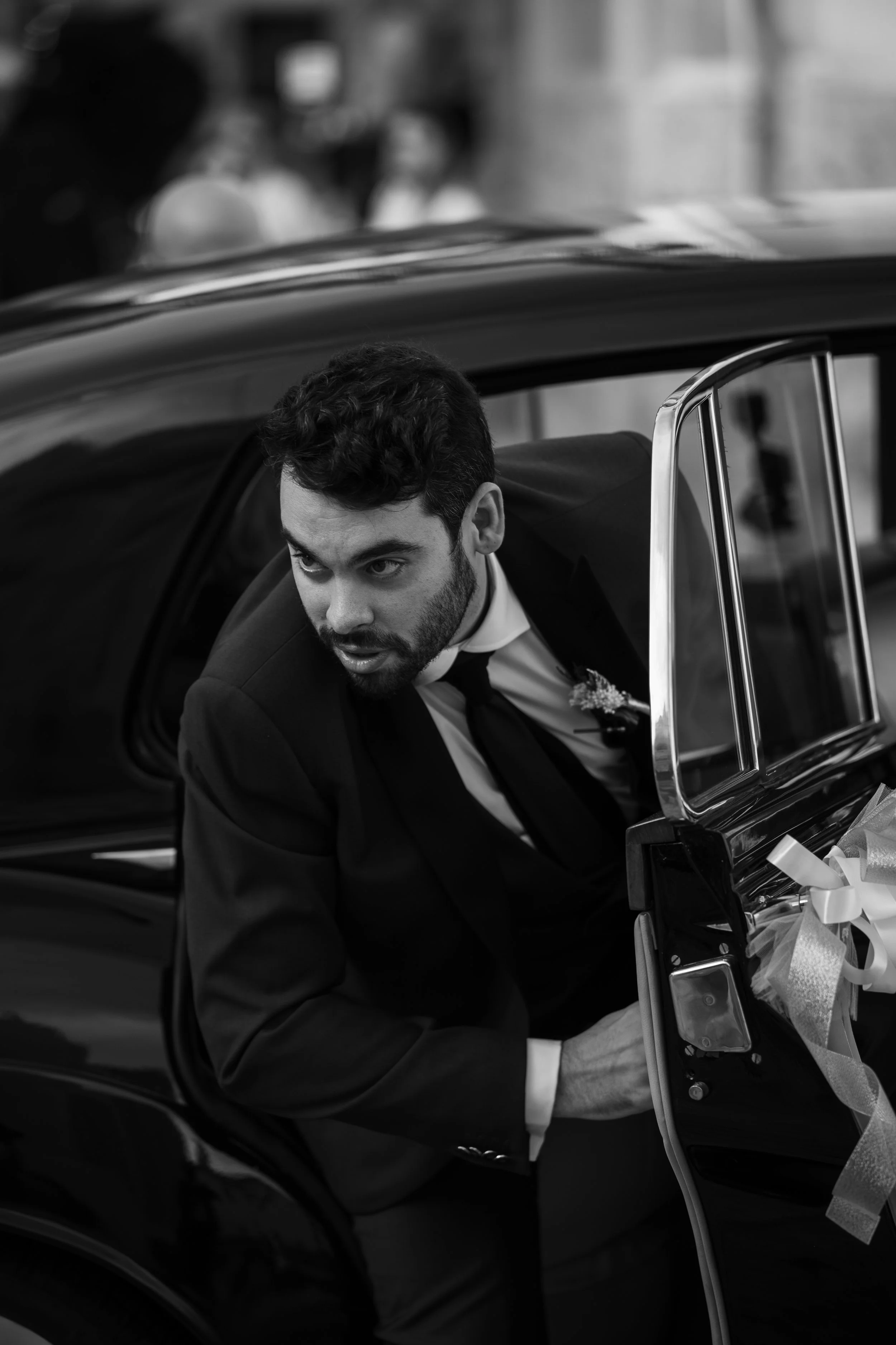 Hombre vestido con traje negro metiéndose en un automóvil negro. Hay una camilla con una bolsa de medicinas y cinta en la parte superior derecha del marco.