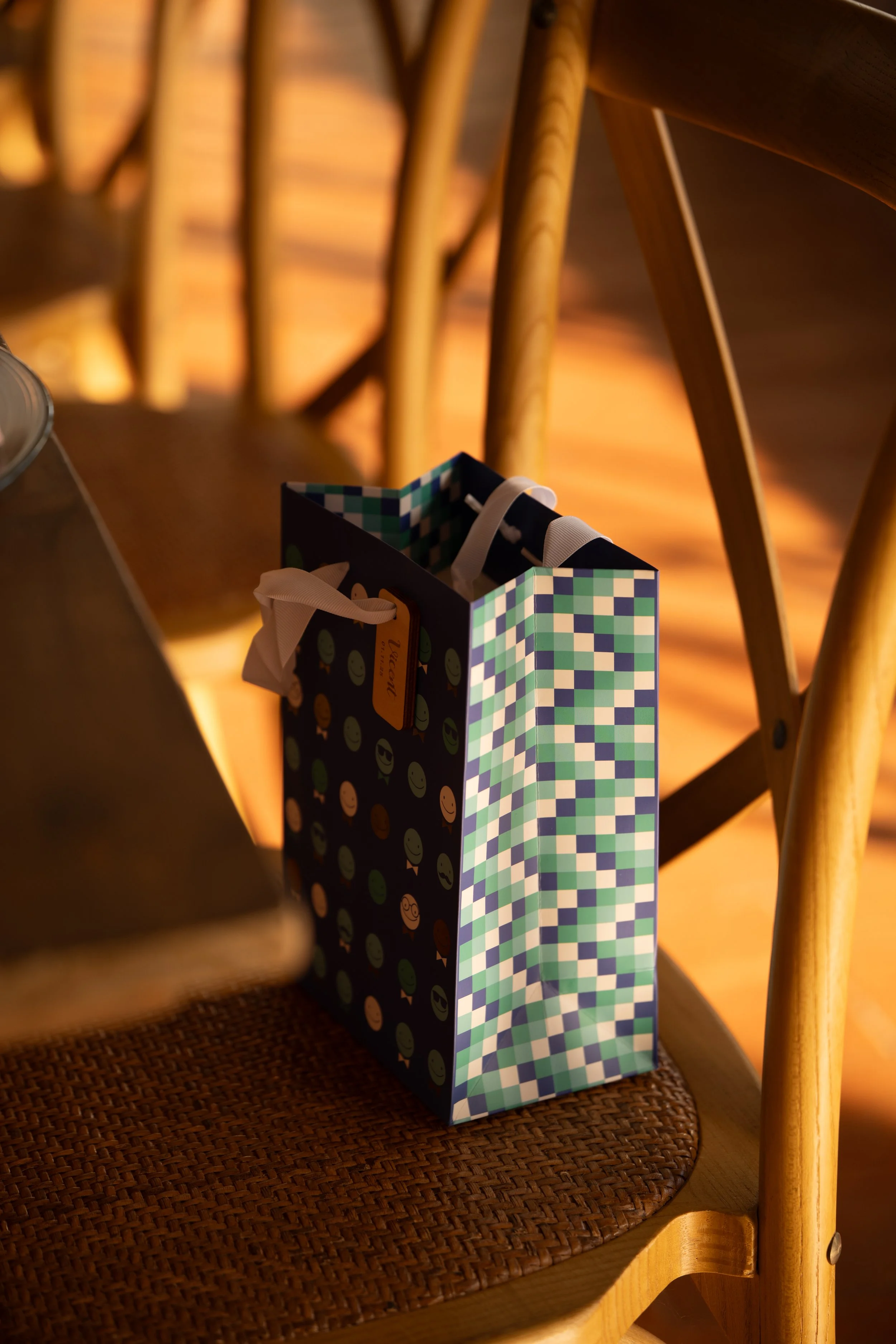Una bolsa de regalo con diseño de cuadrados en tonos verdes y azules, colgada de una silla de madera en un ambiente cálido y bien iluminado.
