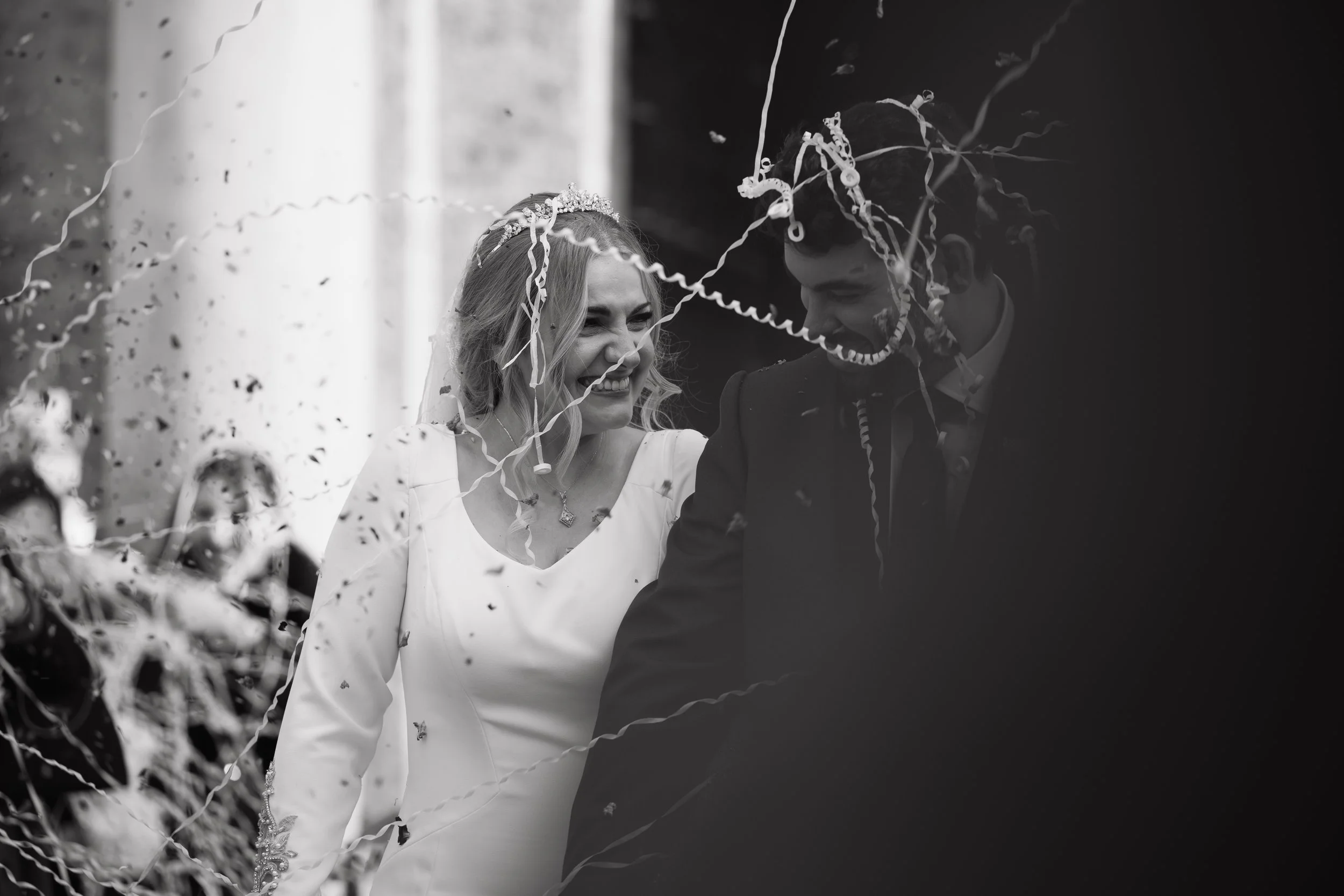 Una pareja de novios sonrientes en su boda, rodeados de serpentinas y confeti, compartiendo un momento feliz.