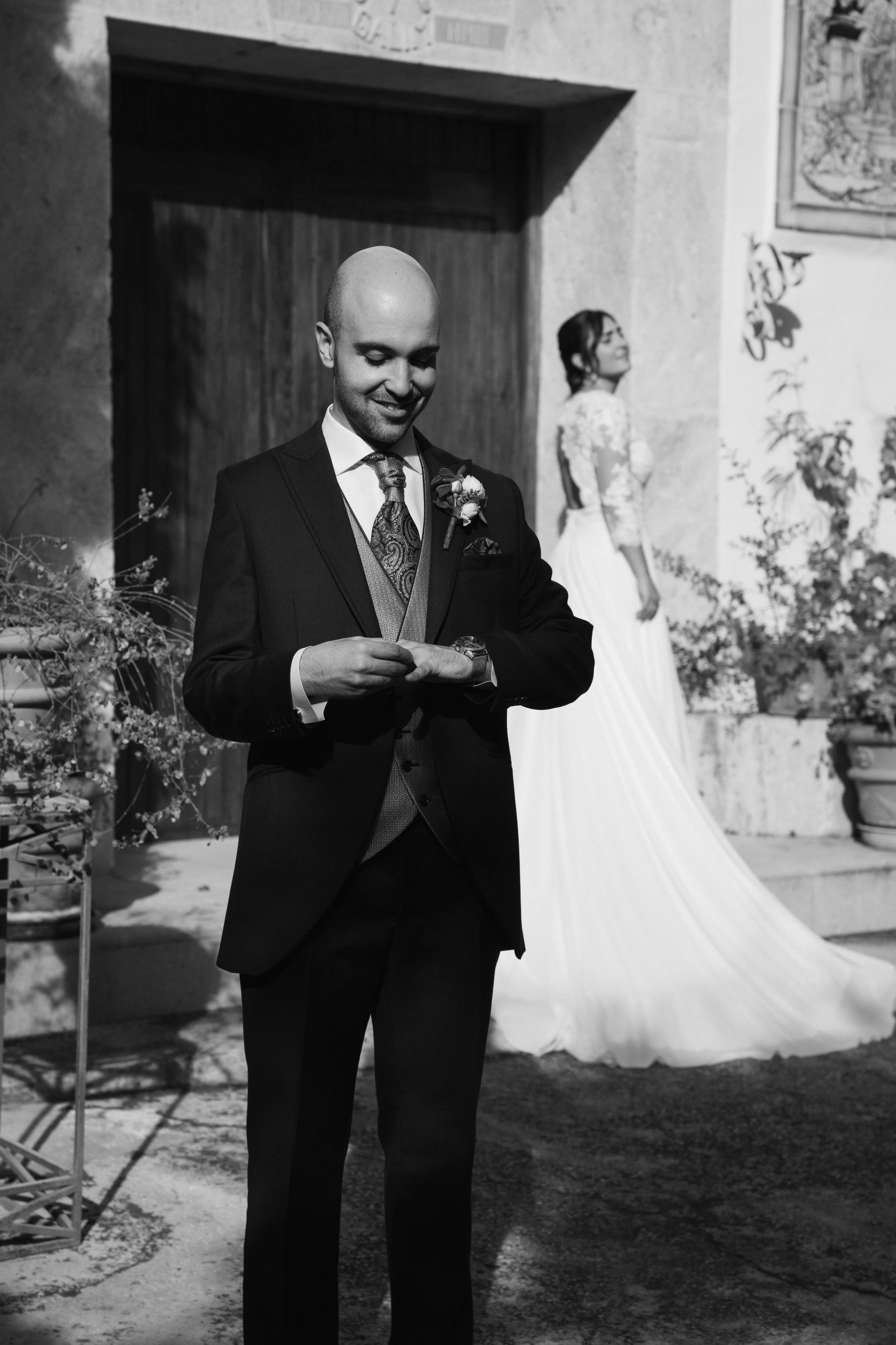 Hombre con traje y corbata en una boda, sonriente y ajustándose la pulsera, con una mujer vestida de novia al fondo, en un lugar decorado con plantas y cuadros.