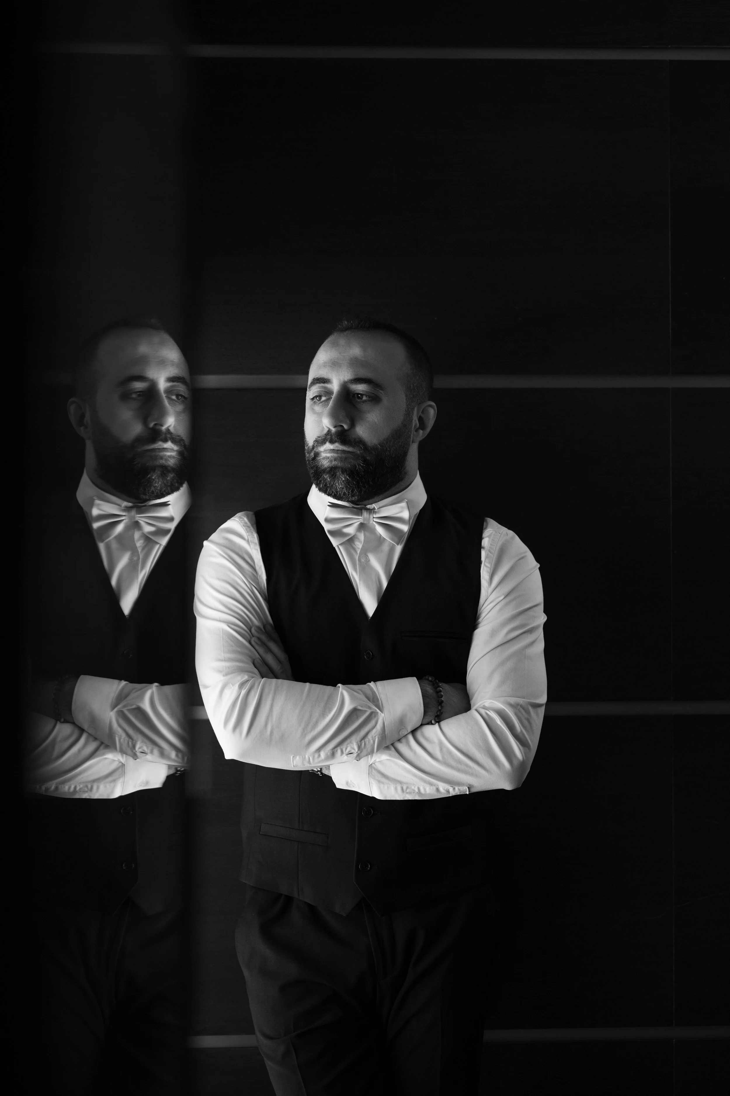 Hombre con barba y cabello corto, vestido con camisa blanca, chaleco y pajarita, mirando hacia la derecha, reflejado en un espejo en un entorno oscuro.