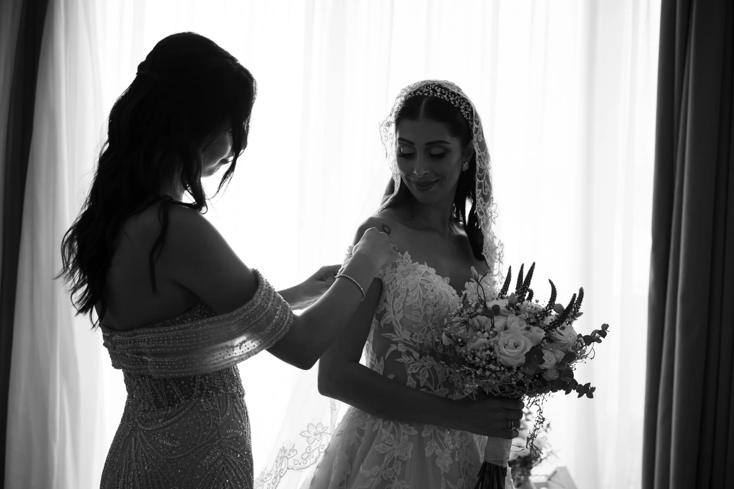 Mujer en vestido de boda recibiendo un ramo de flores, con otra mujer ayudándola, en un entorno con cortinas blancas de fondo, en una ceremonia de boda.