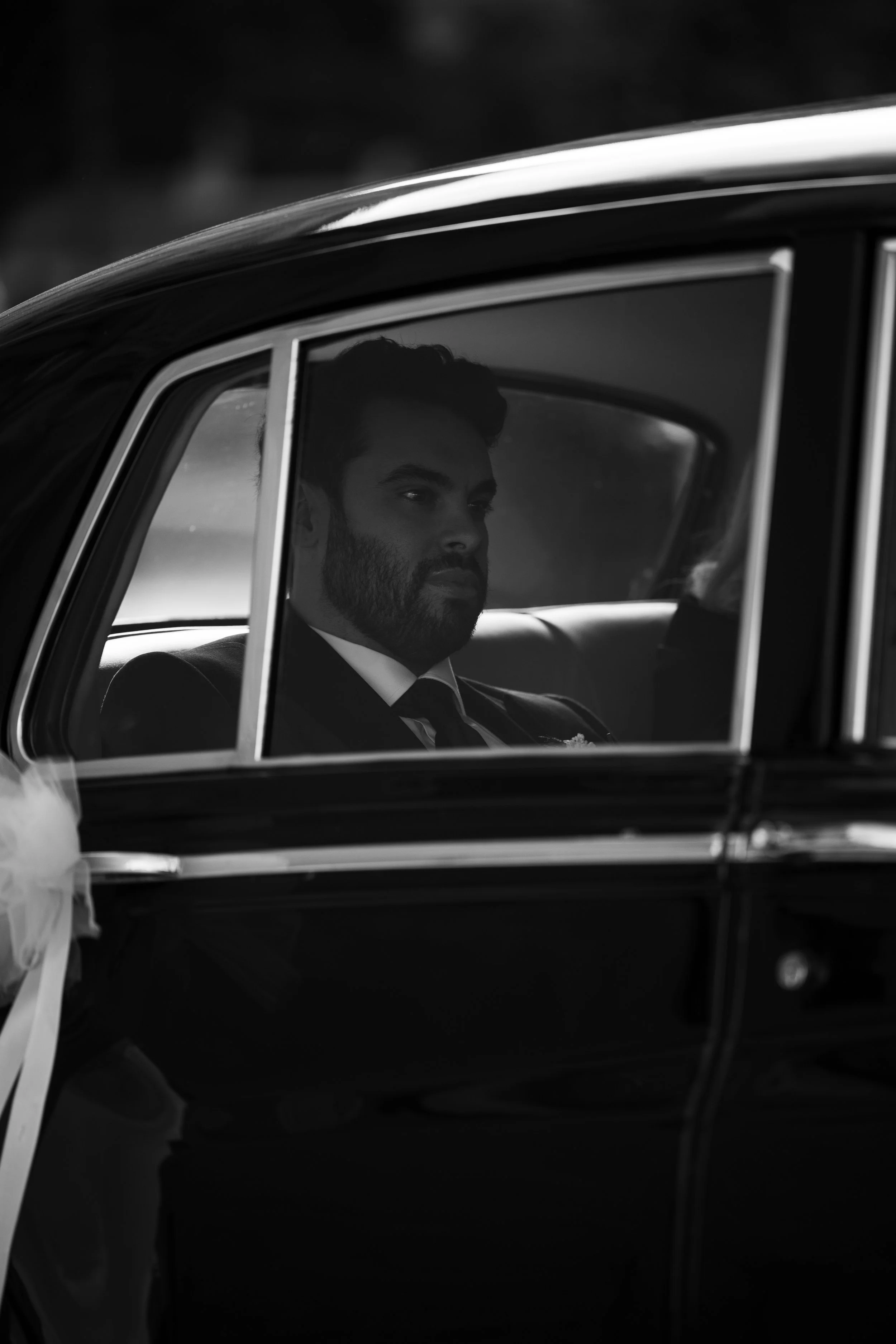 Hombre con barba y bigote, vestido formalmente, sentado en un coche clásico, en un ambiente en blanco y negro.