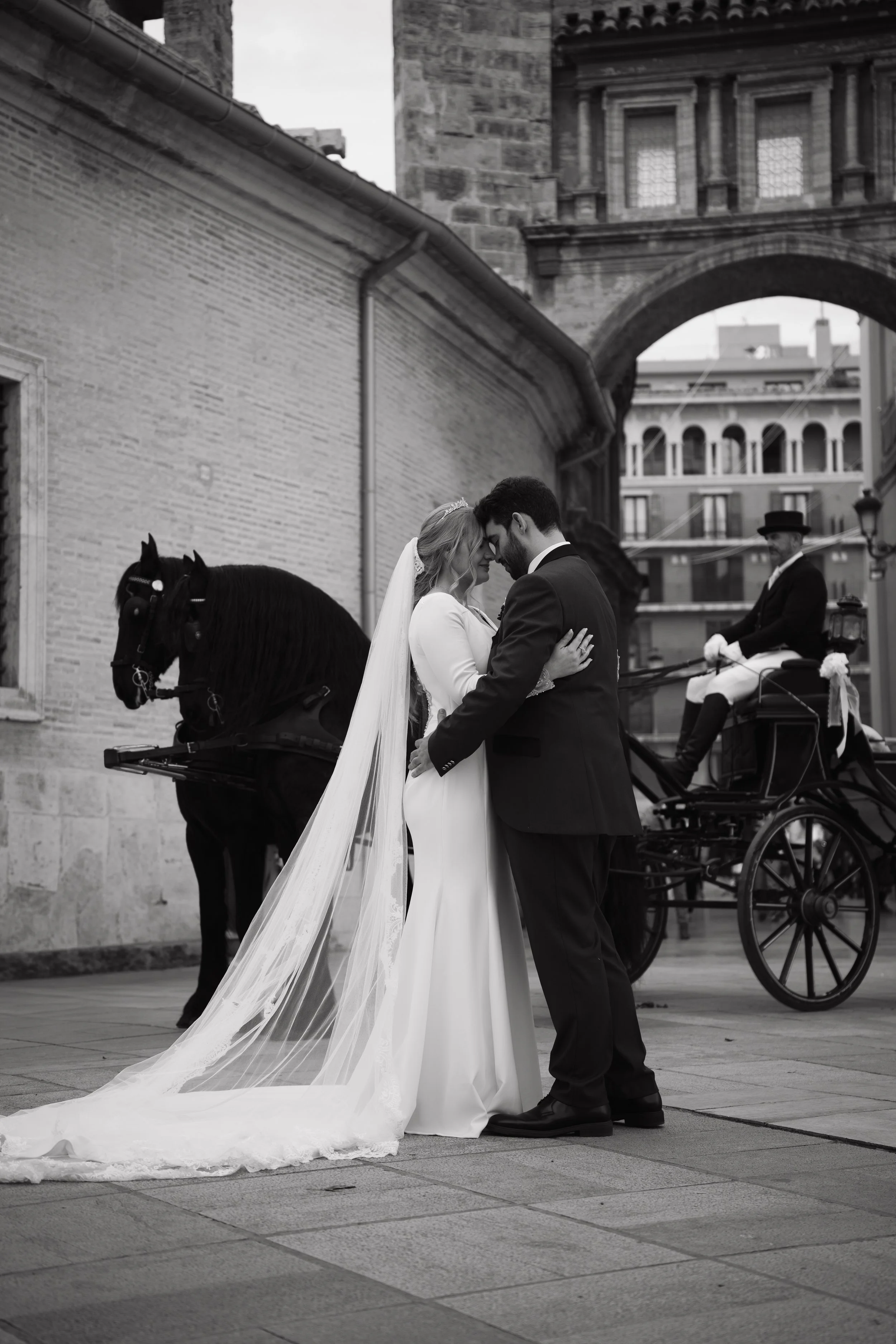 Pareja de novios en la calle, la novia viste vestido largo de boda y el novio traje oscuro, ambos se miran y se tocan la frente, en el fondo hay una carreta con caballos y un hombre con sombrero, en un entorno urbano con edificios históricos.
