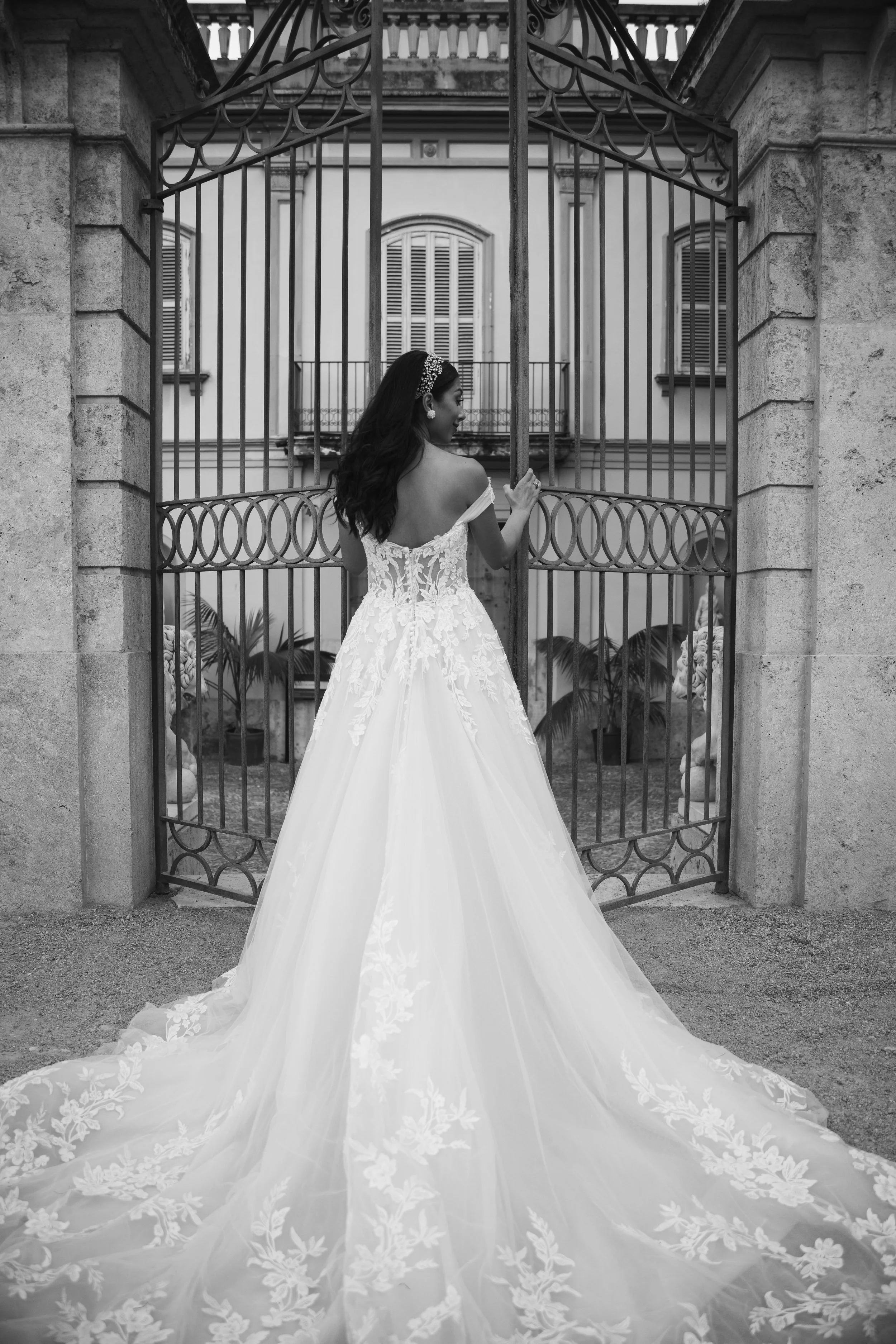 Mujer en vestido de novia de espaldas, con un arco y rejas de hierro forjado, en una escena en blanco y negro, frente a un edificio con ventanas con persianas.