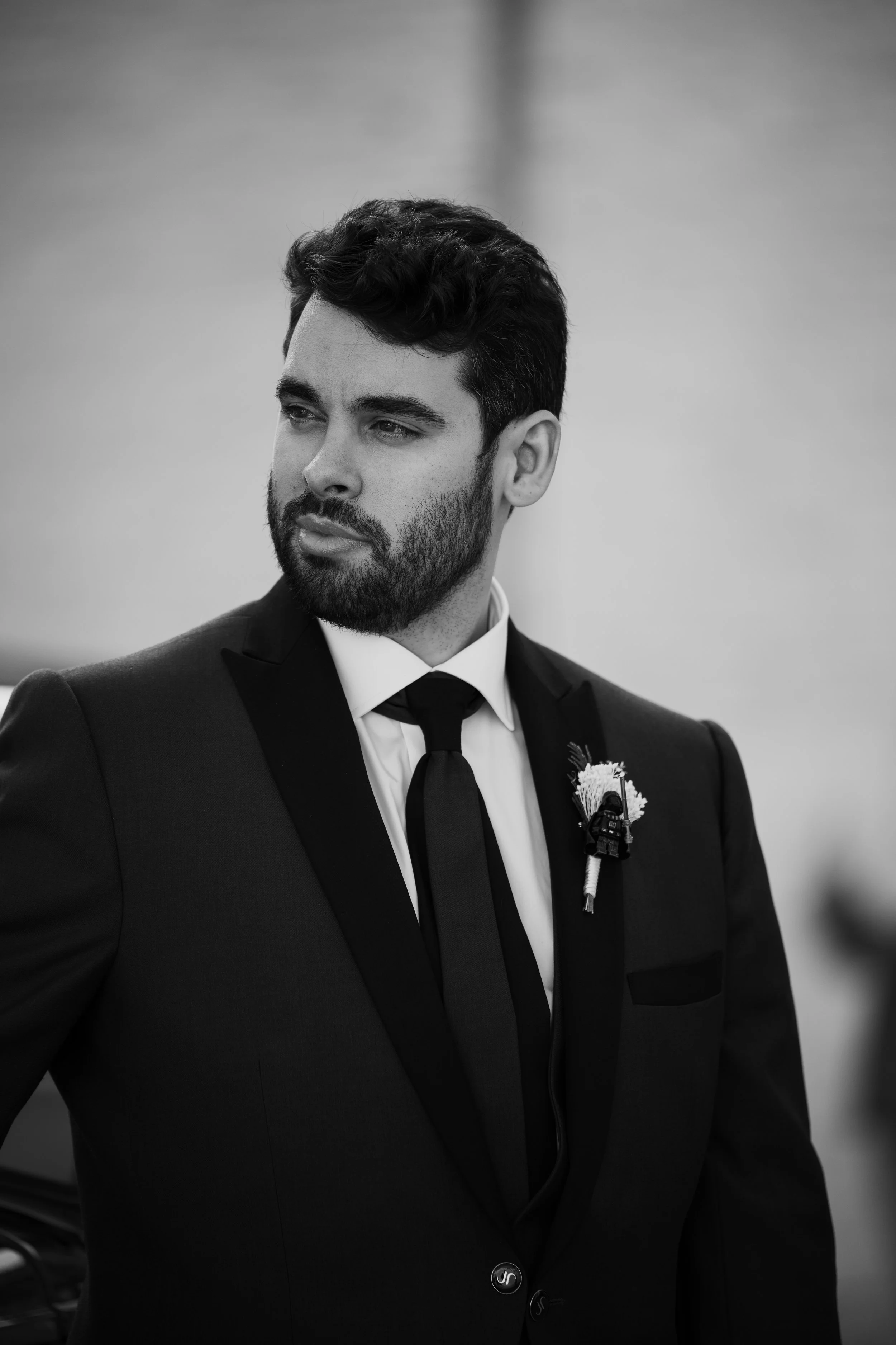 Hombre con barba y cabello oscuro, veste un traje negro con camisa blanca y corbata negra, lleva un bouton de flor y un micrófono en el saco, en un fondo difuso.