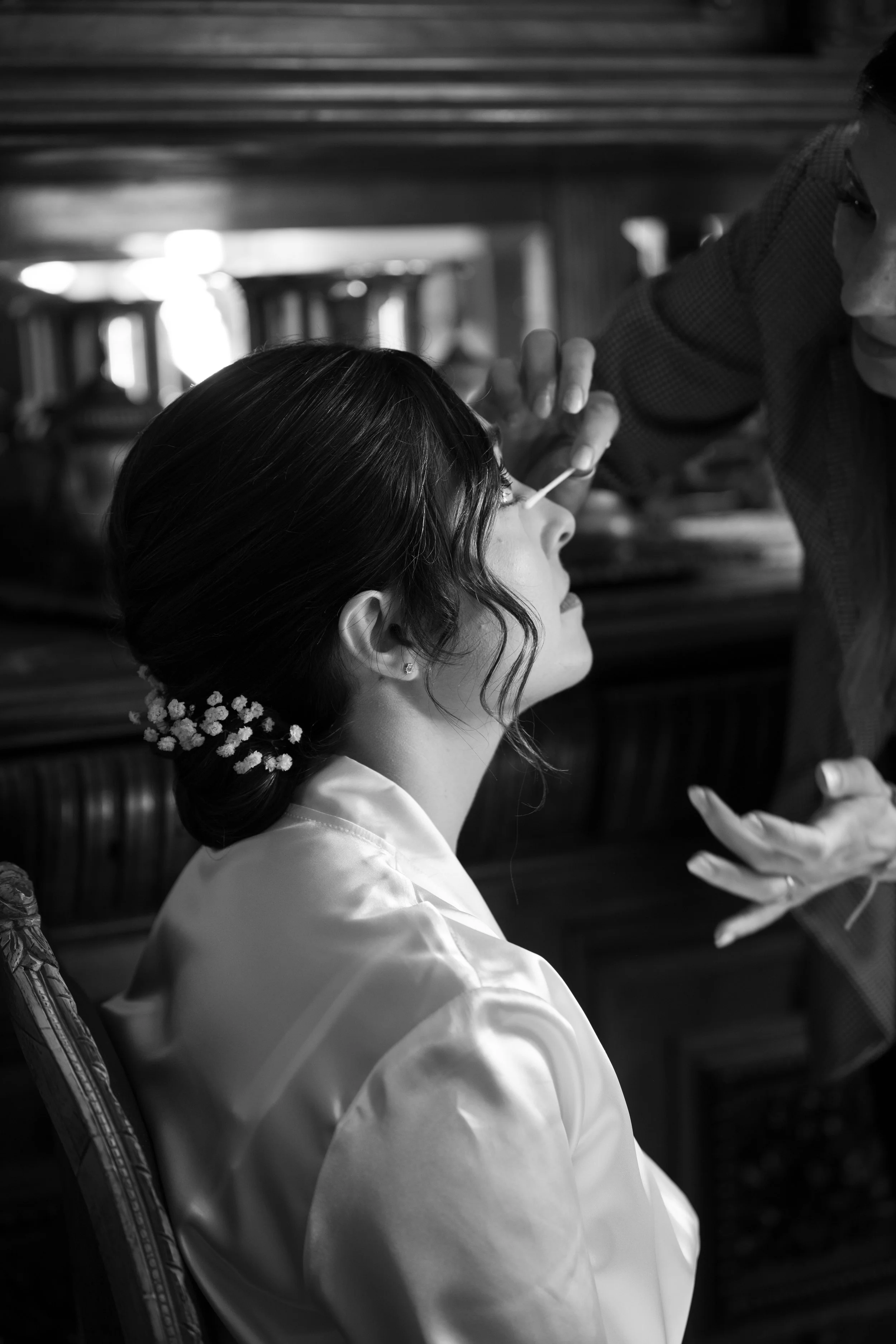 Mujer con cabello oscuro recogido decorado con flores, vestida con ropa blanca, sentada en una silla, en un entorno que parece ser un lugar cerrado, mientras una persona la atiende, posiblemente para un procedimiento de maquillaje o cuidado personal.