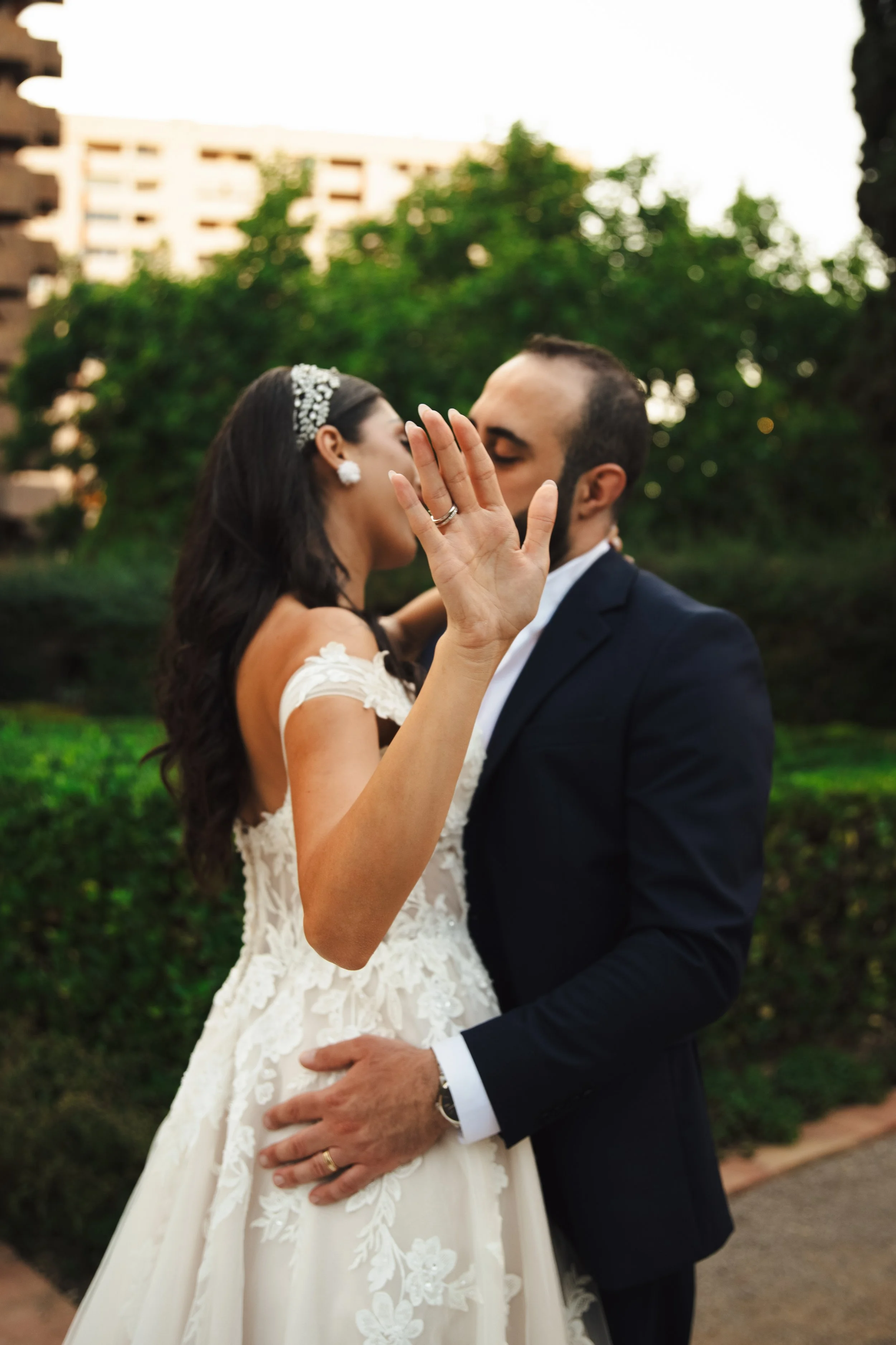 Pareja de recién casados en un beso romántico en un parque con árboles y edificios de fondo.