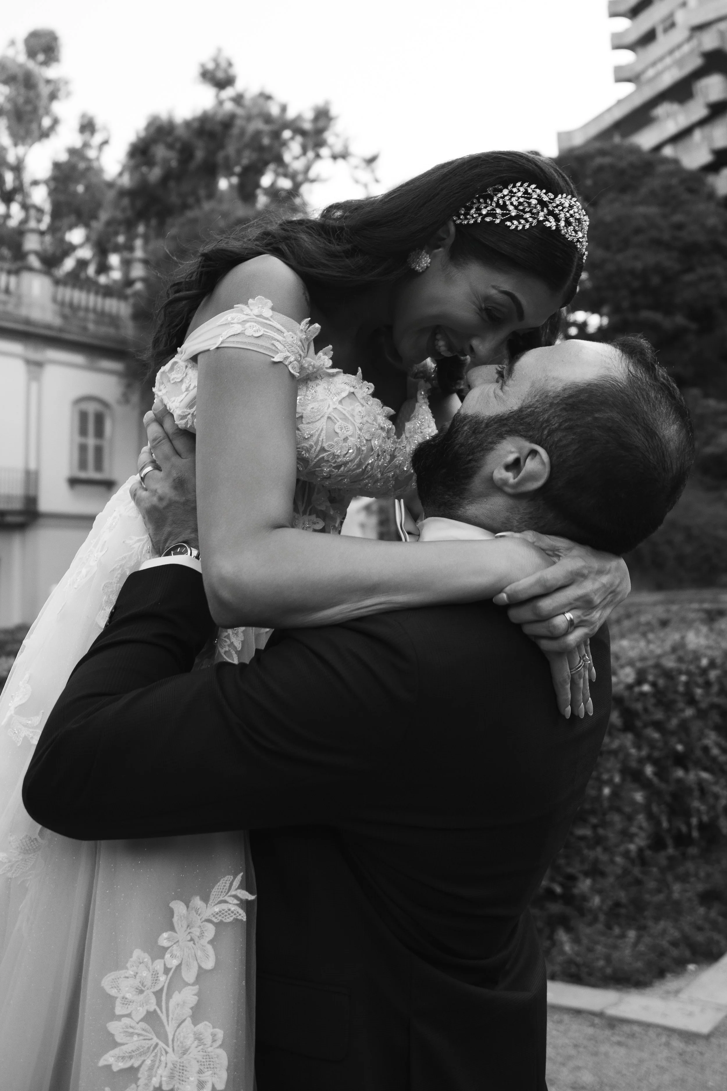 Una pareja en boda, el hombre sostiene a la mujer en sus brazos y ambos se ven felices, en un entorno exterior con árboles y un edificio en el fondo.
