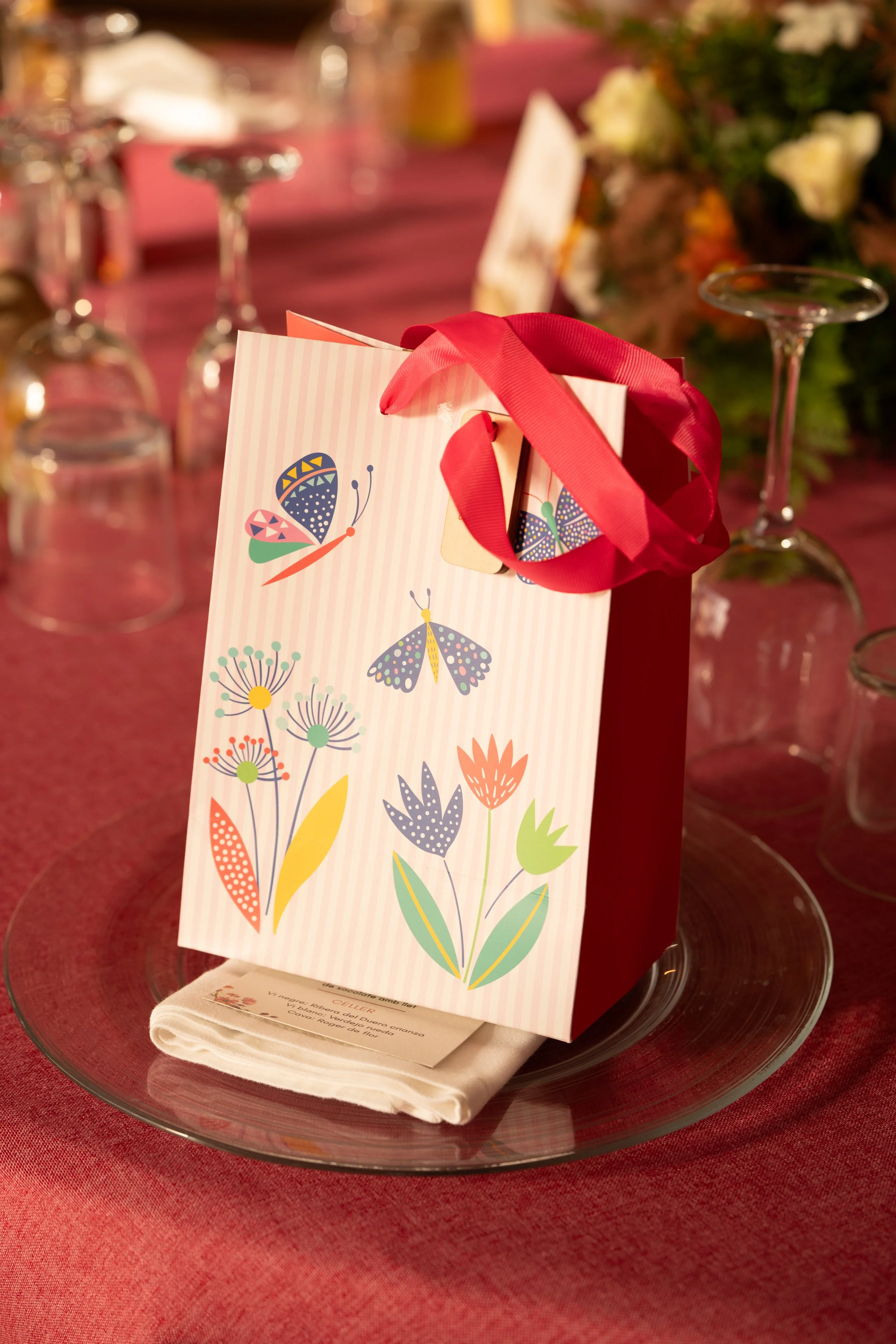 Sobre de regalo con dibujos de mariposas y flores, rodeado de copas de vino invertidas en una mesa con mantel rojo