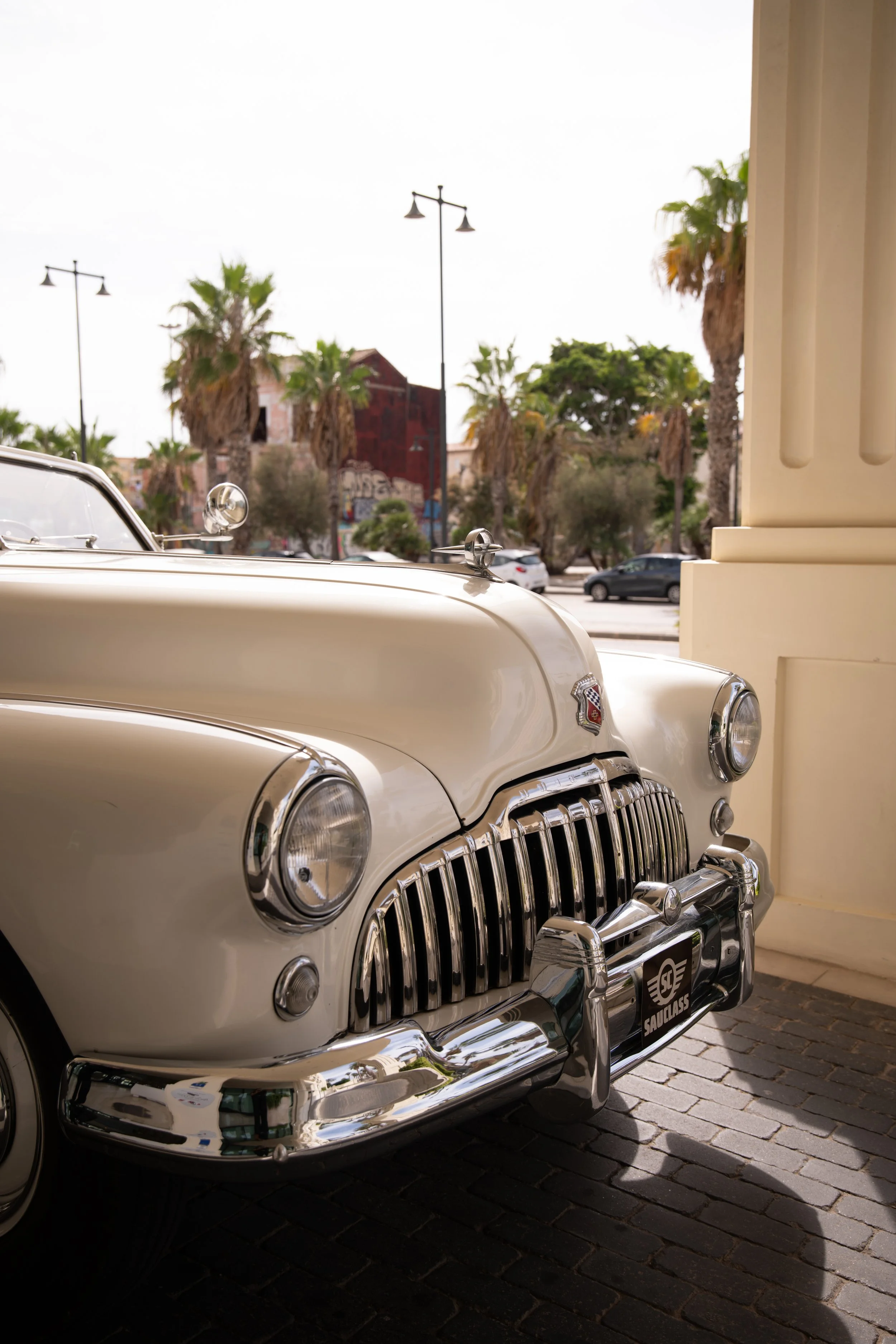 Primer plano de un coche clásico blanco, parcialmente cubierto por sombra, con un diseño de parrilla y faros antiguos y un emblema en el capó.