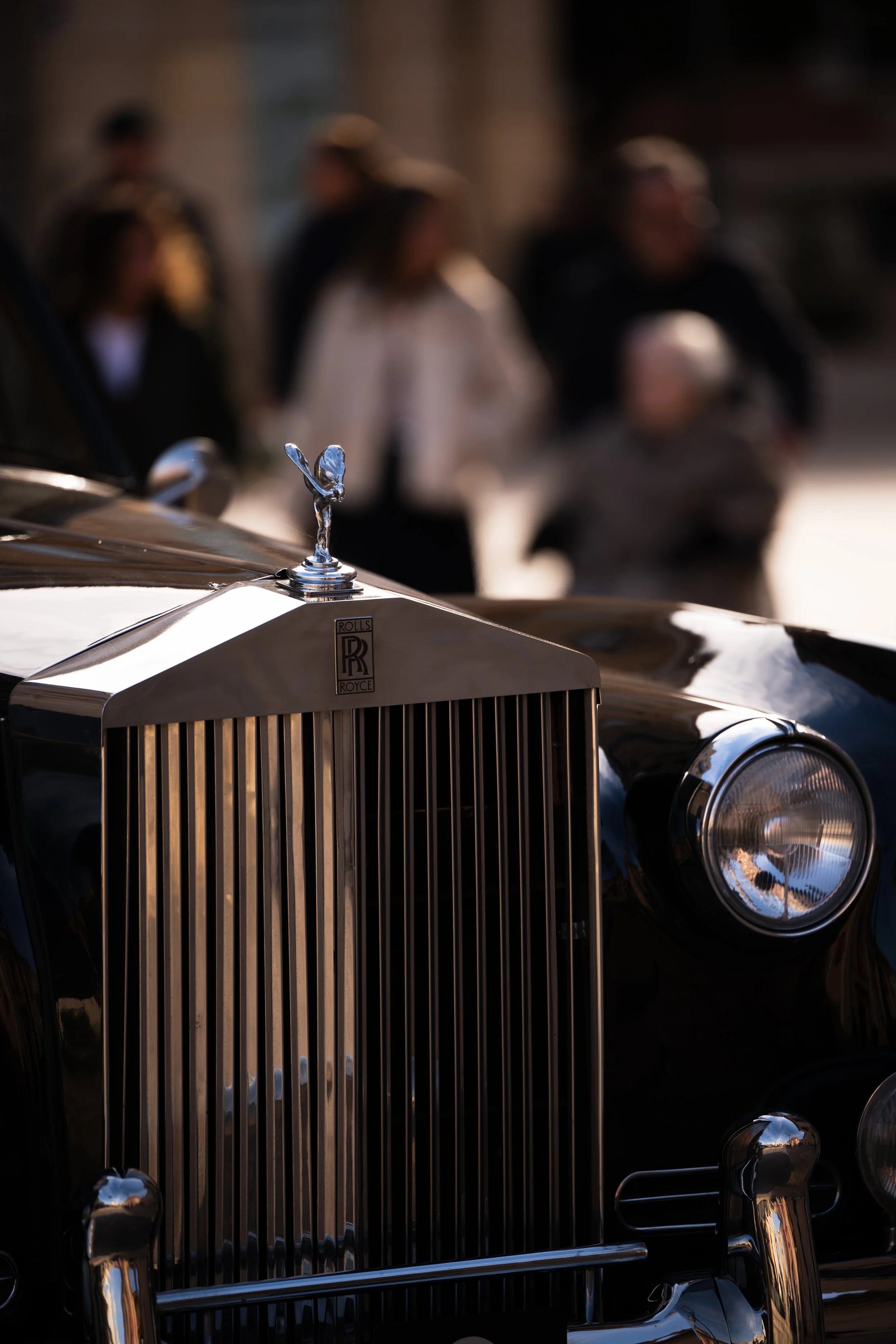 Primer plano de una parrilla de coche clásico Rolls Royce con el emblema Spirit of Ecstasy en la parte superior del radiador. En el fondo, personas desplazándose por la calle, desenfocadas.