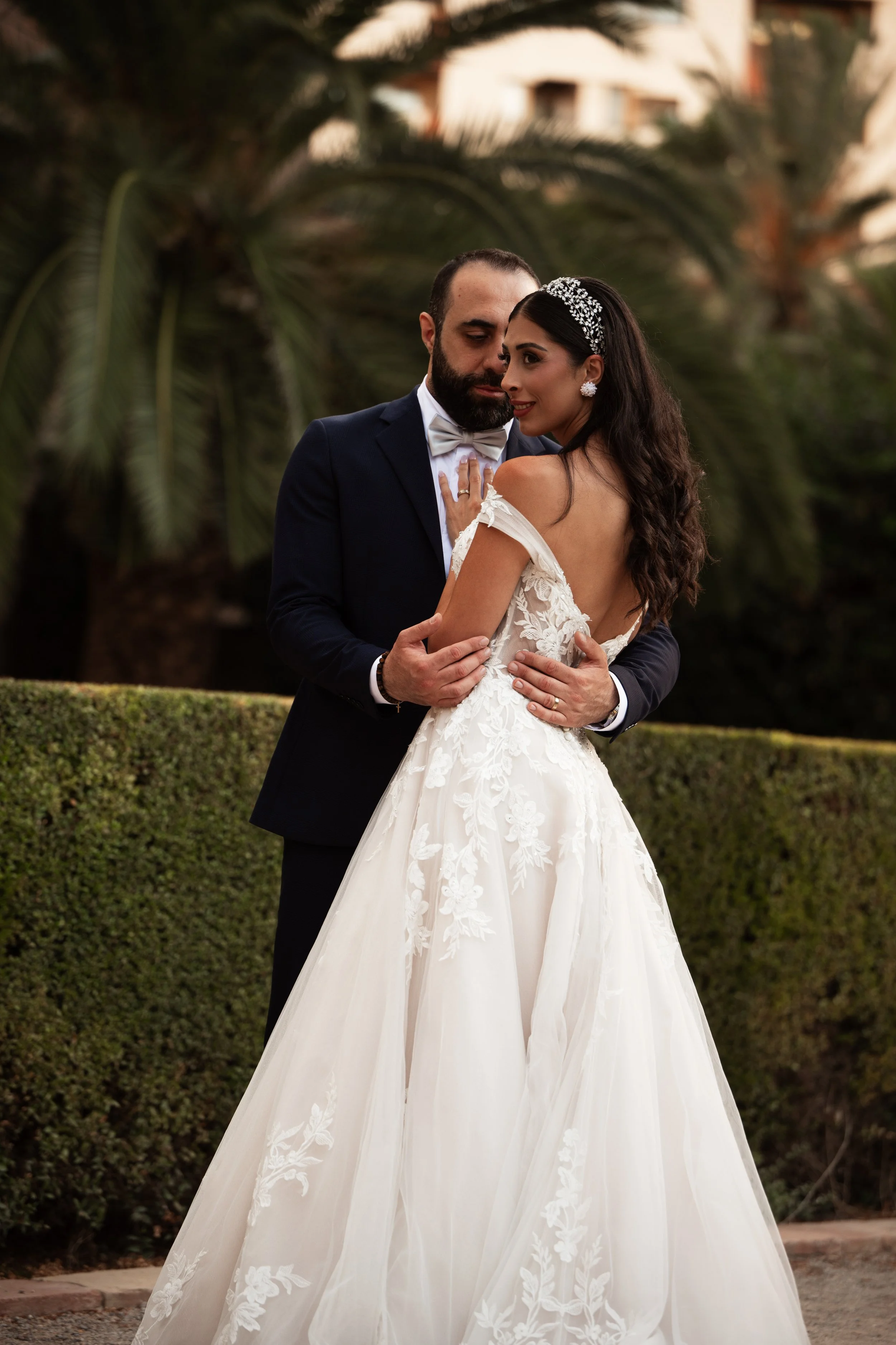 Pareja de recién casados abrazados en un jardín con árboles en el fondo, ella lleva vestido de novia y él traje de novio.