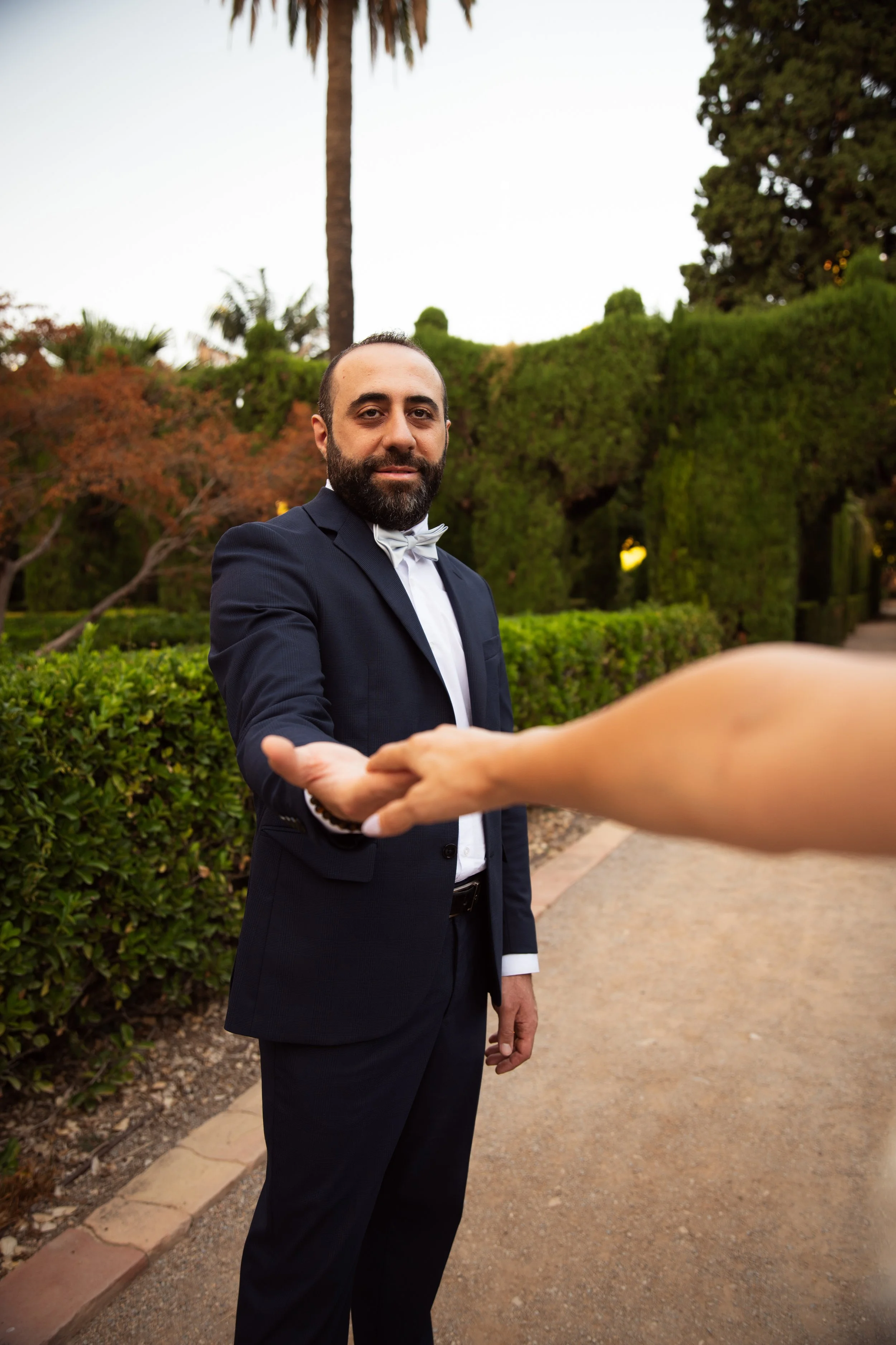 Un hombre con barba, vestido con traje formal y una pajarita, extendiendo su mano para estrechar la mano de otra persona en un jardín con árboles y arbustos verdes, al atardecer.