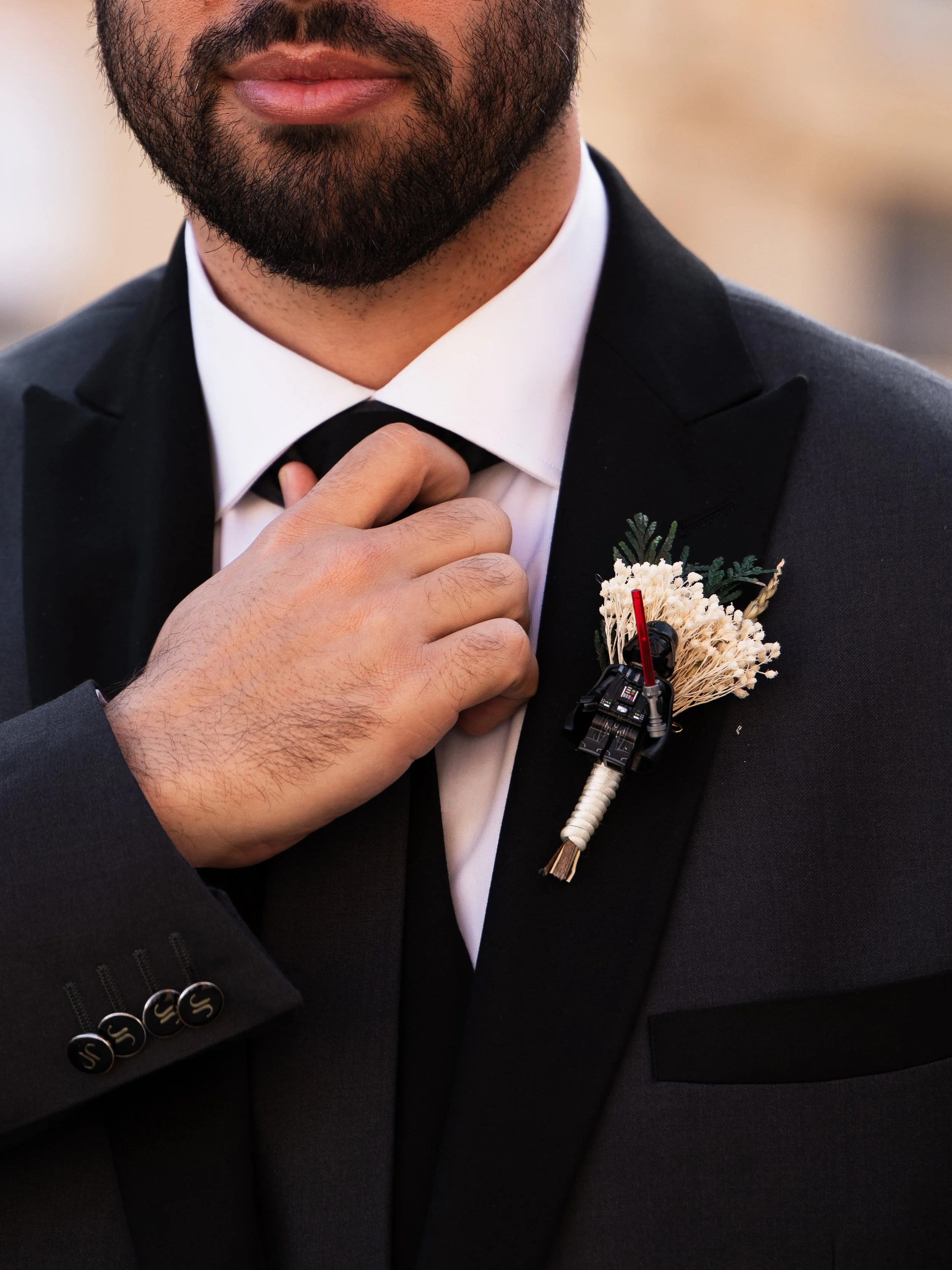 Hombre con barba arreglada, vestido con traje negro y corbata, ajustándose la camisa en un evento formal, con flor y un micrófono en el saco.