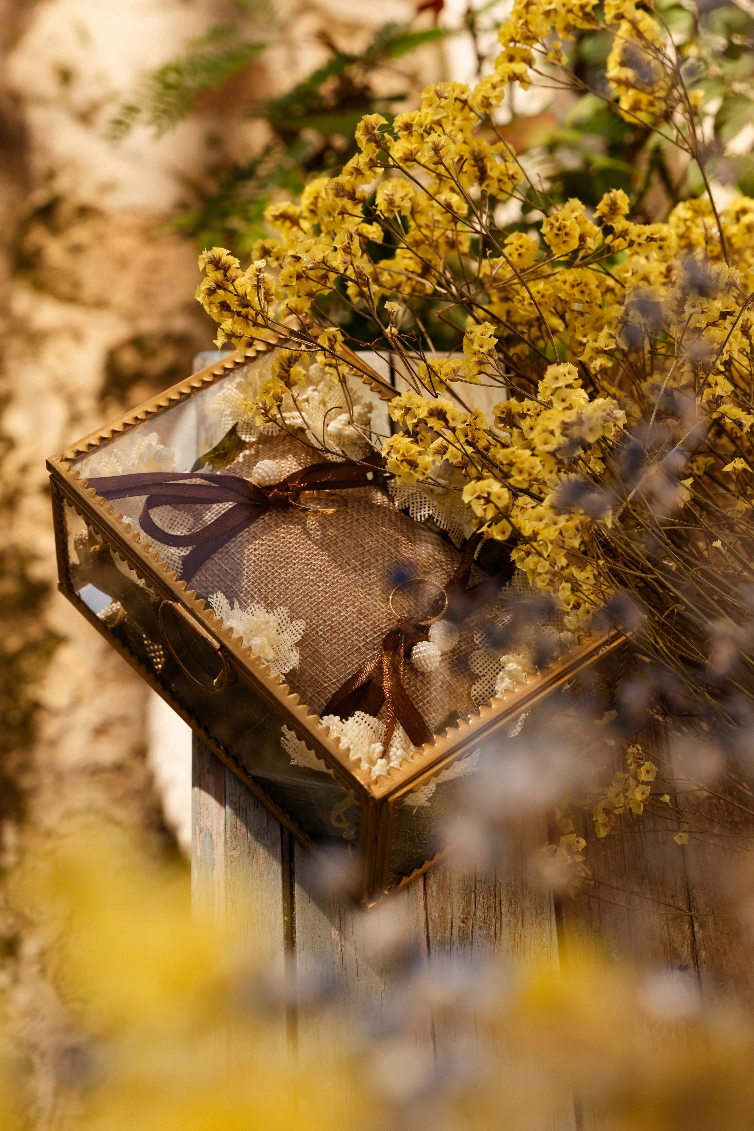 Una caja decorativa con tela y listón de color marrón, que contiene anillos de boda y decoraciones blancas. Encima hay un ramo de flores amarillas.