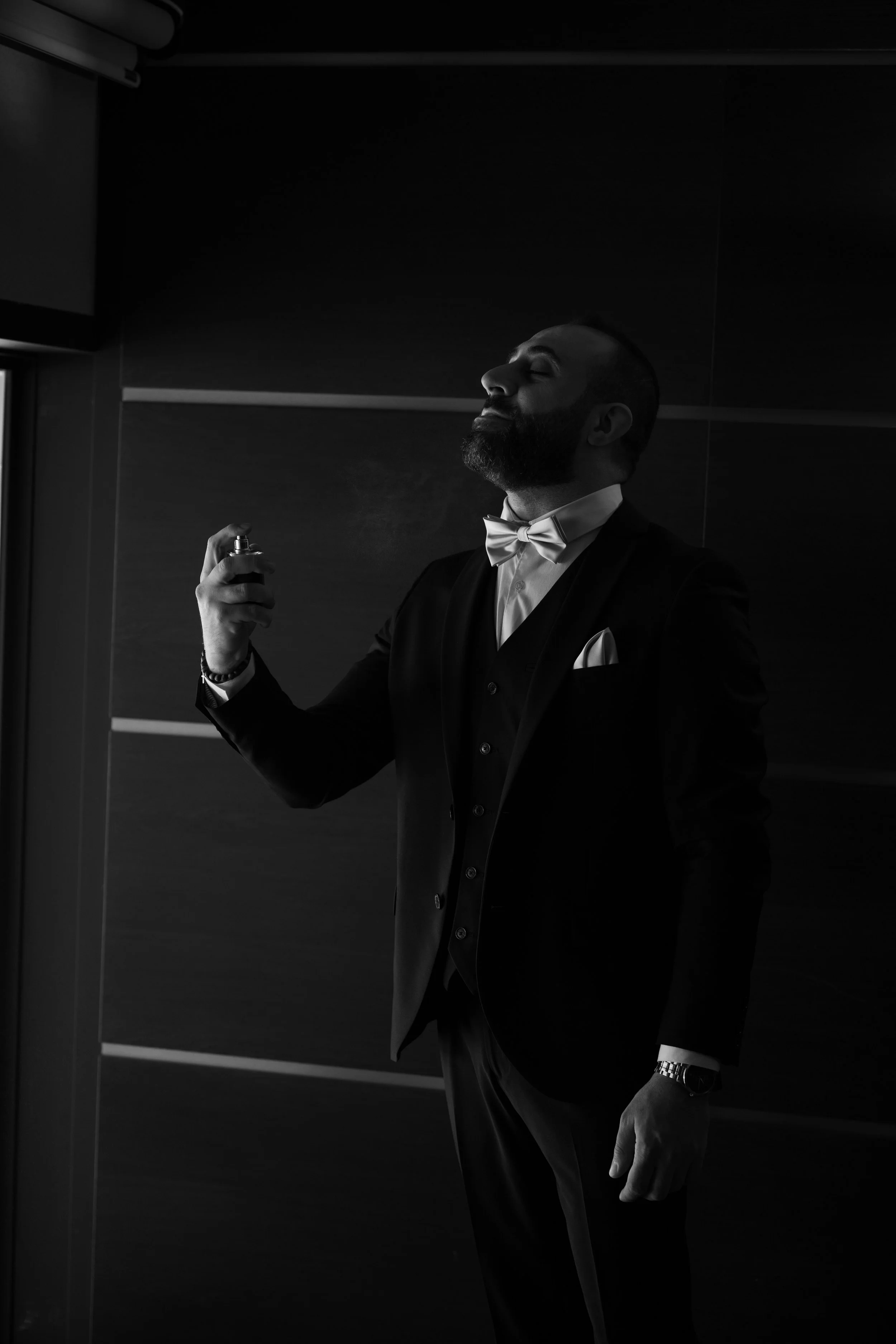 Hombre con barba y traje ajustado, con pajarita blanca, rociando perfume, en un ambiente con paredes de madera, en un estilo en blanco y negro.