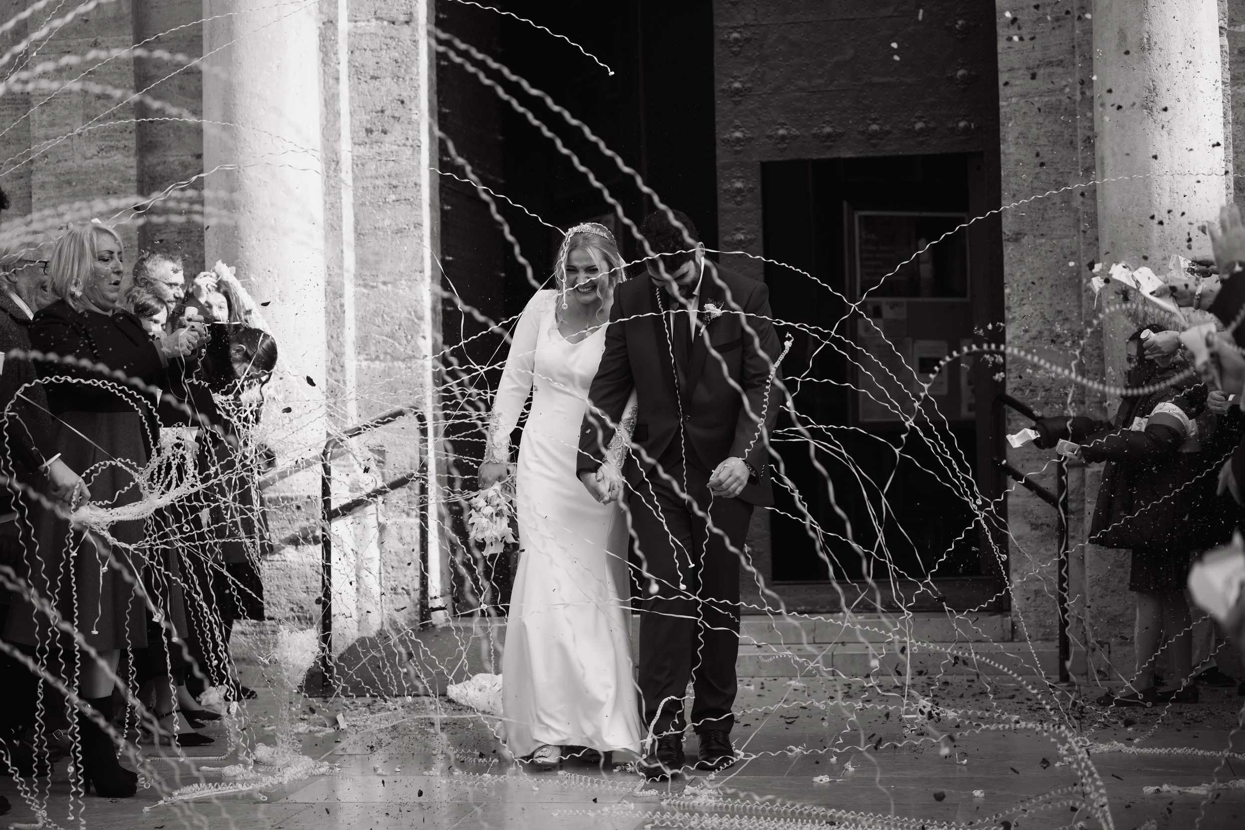 Pareja de recién casados en su boda, caminando de la mano después de la ceremonia, rodeados de invitados lanzando serpentinas.