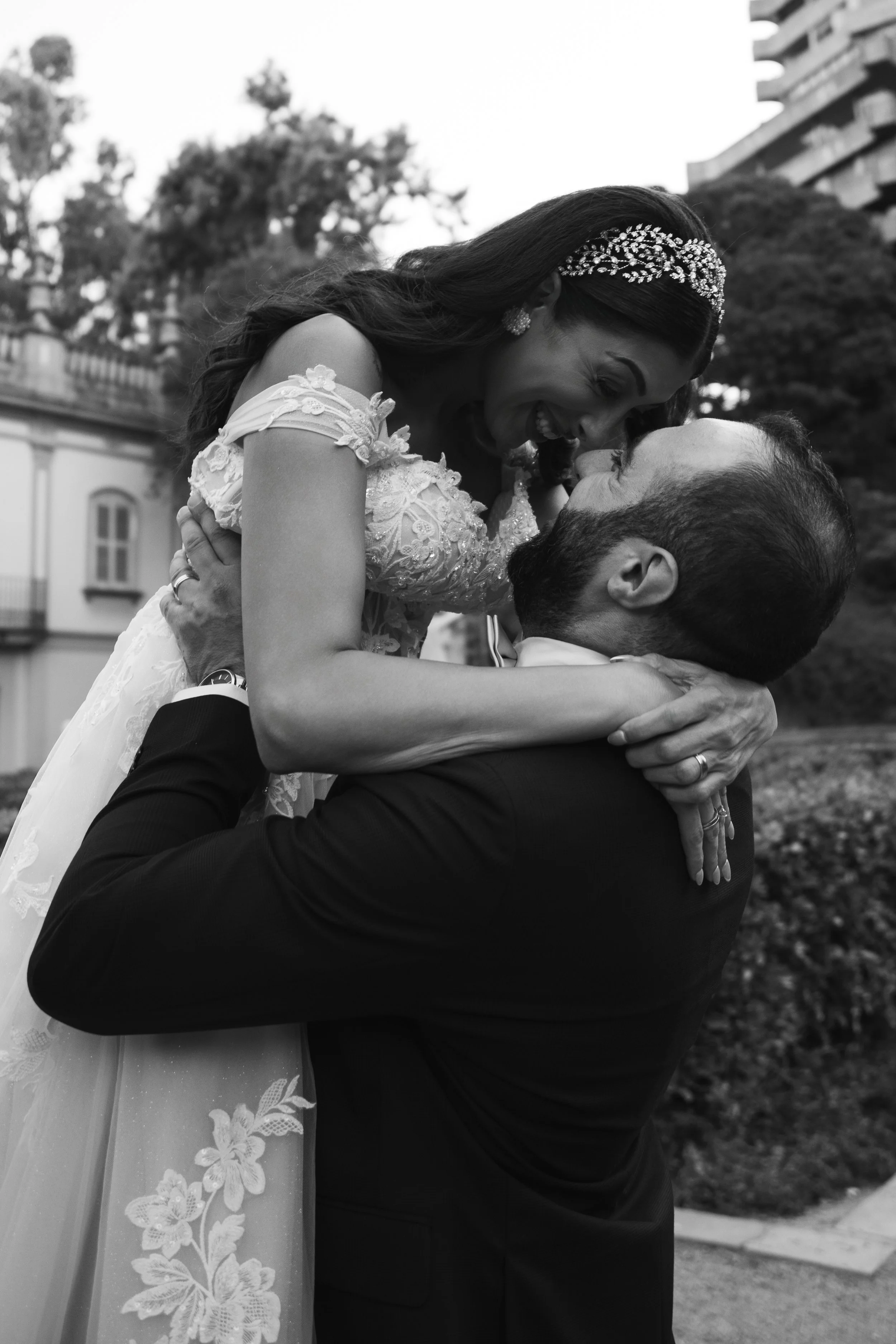 Boda de una pareja feliz, ella lleva vestido y diadema y él viste traje negro, se abrazan y se miran con alegría en un entorno exterior.