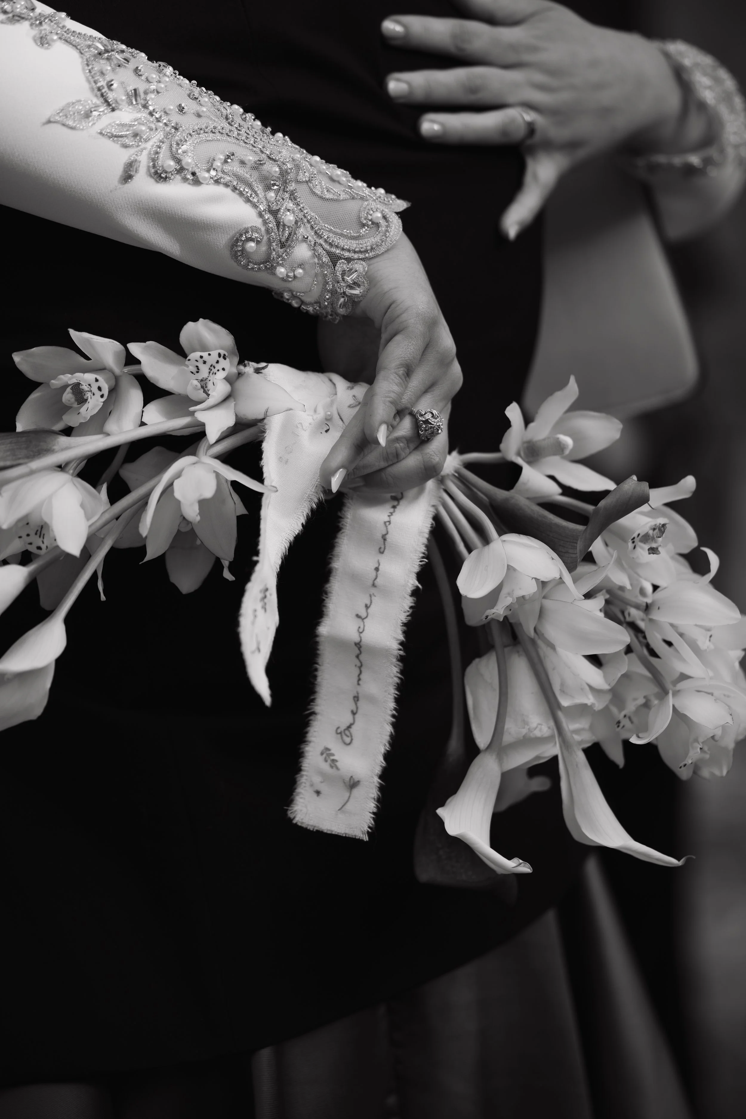 Manos con anillos y vestido de novia con bordados, sosteniendo un ramo de flores de orquídeas blancas y cinta con escritura en letra cursiva.