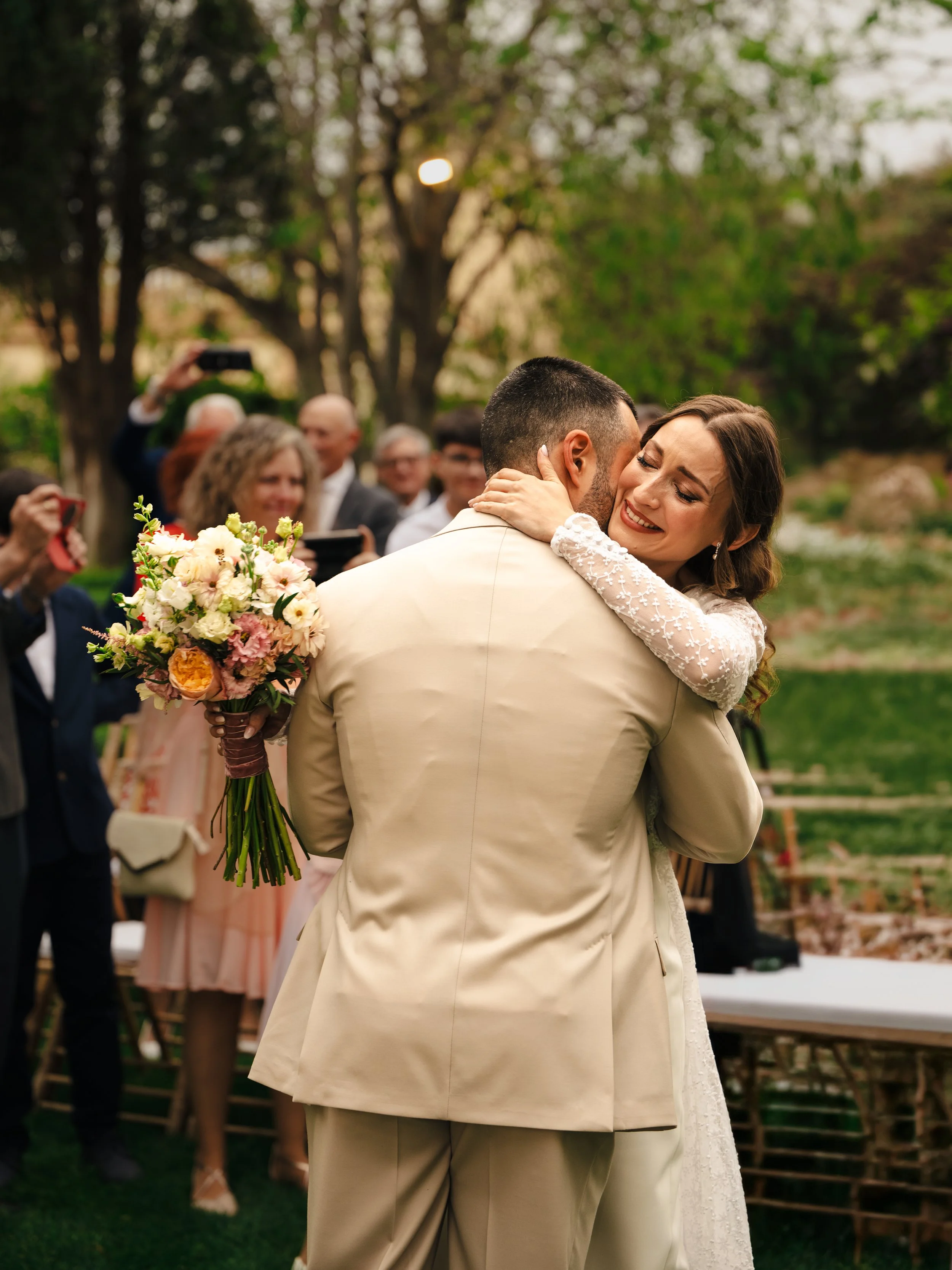 Pareja en vestido de boda abrazados, rodeados de invitados en un jardín, con árboles y un ambiente de celebración.