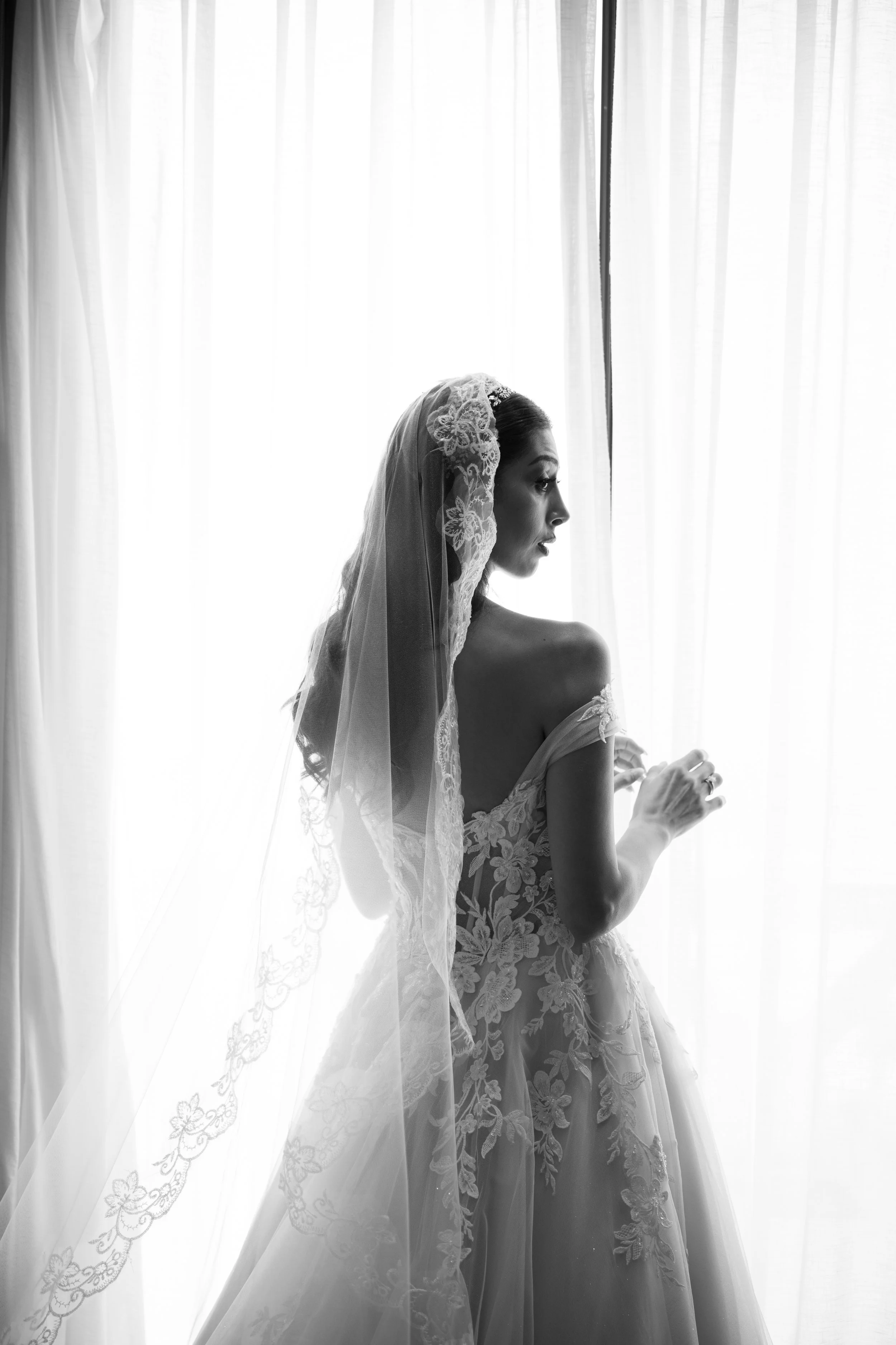 Mujer con vestido de novia y velo, en perfil, frente a cortinas blancas por la ventana.